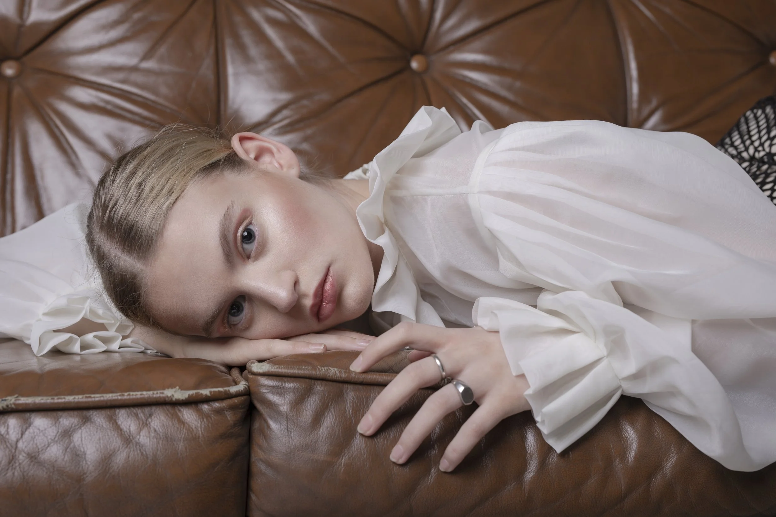 Young woman lying on a brown leather couch, resting her head on her hand, wearing a white blouse and looking at the camera.