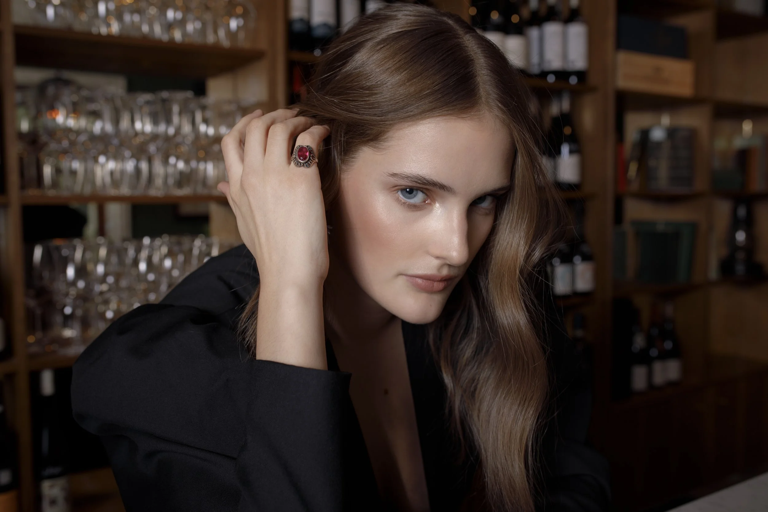 A young woman with long, wavy brown hair and blue eyes, wearing a black top, is resting her head on her hand with a ring on her finger. She is in a bar or restaurant setting with shelves of glasses and bottles in the background.