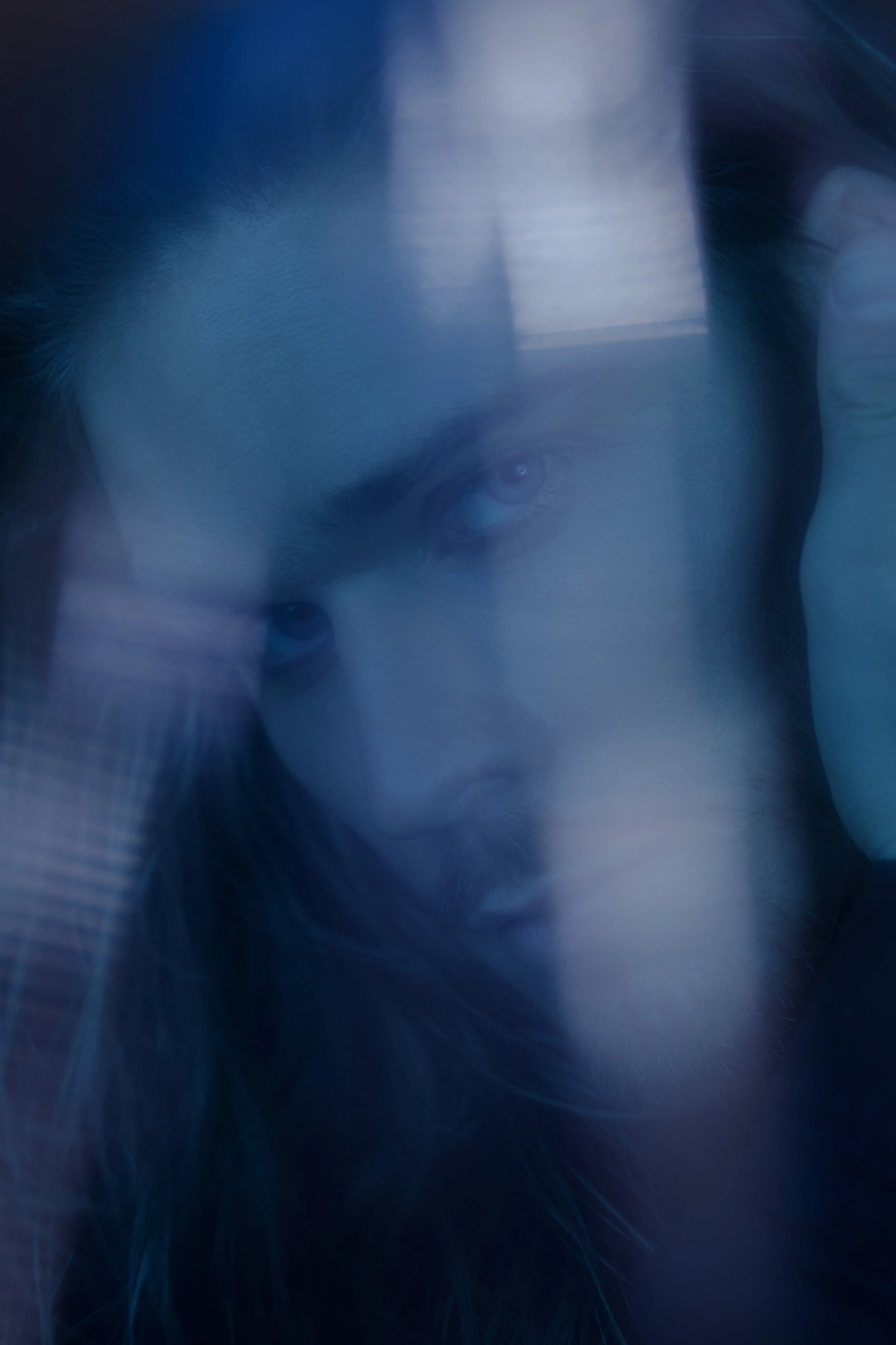 Close-up of a man with long hair, looking through a glass window with reflections, in dim lighting.