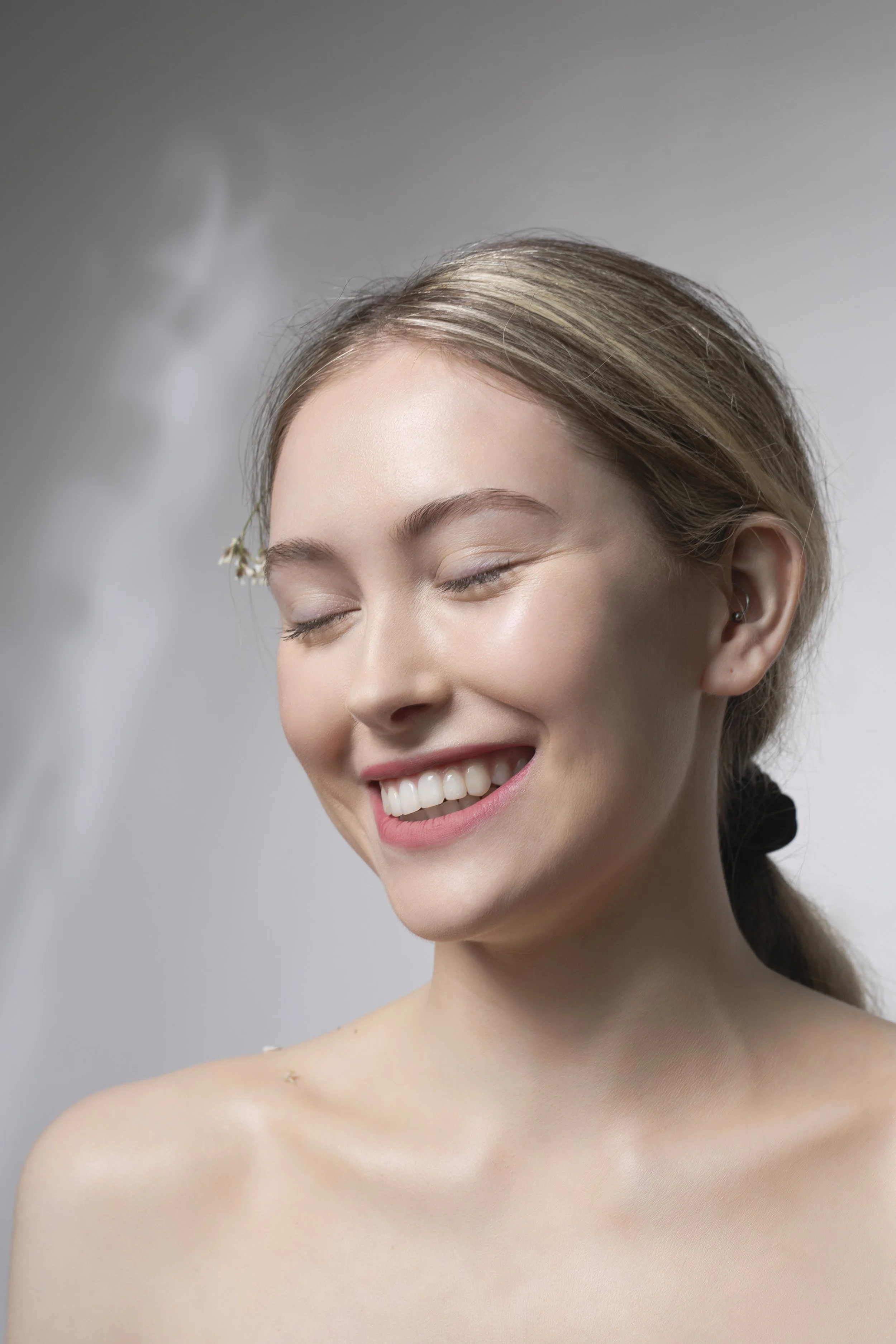 A woman with fair skin and light brown hair tied back, smiling with her eyes closed, showing her teeth, with a soft water spray falling onto her shoulder in the background.