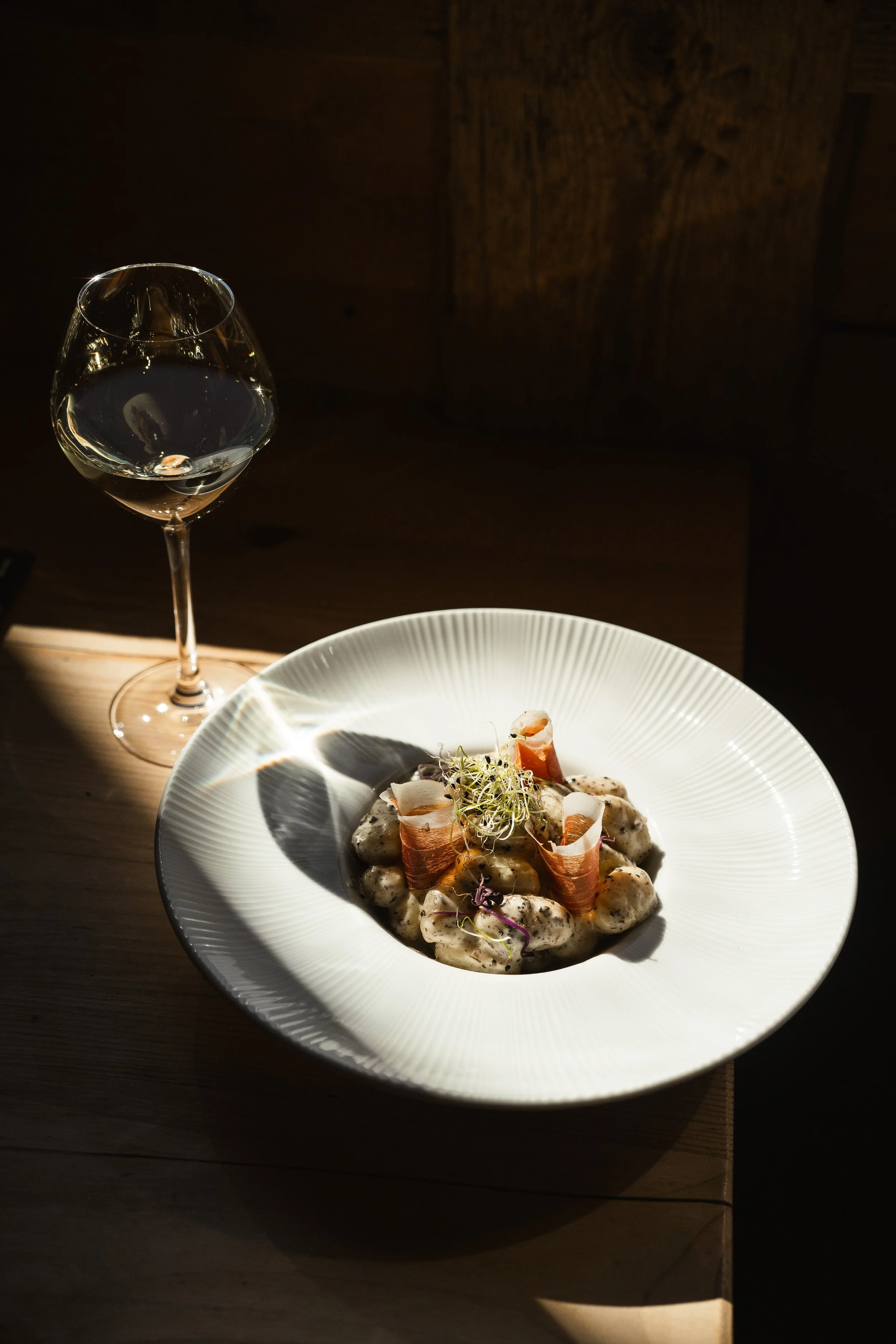 A plate of food with gnocchi covered in creamy sauce, garnished with microgreens and crispy prosciutto, next to a glass of white wine on a wooden table.