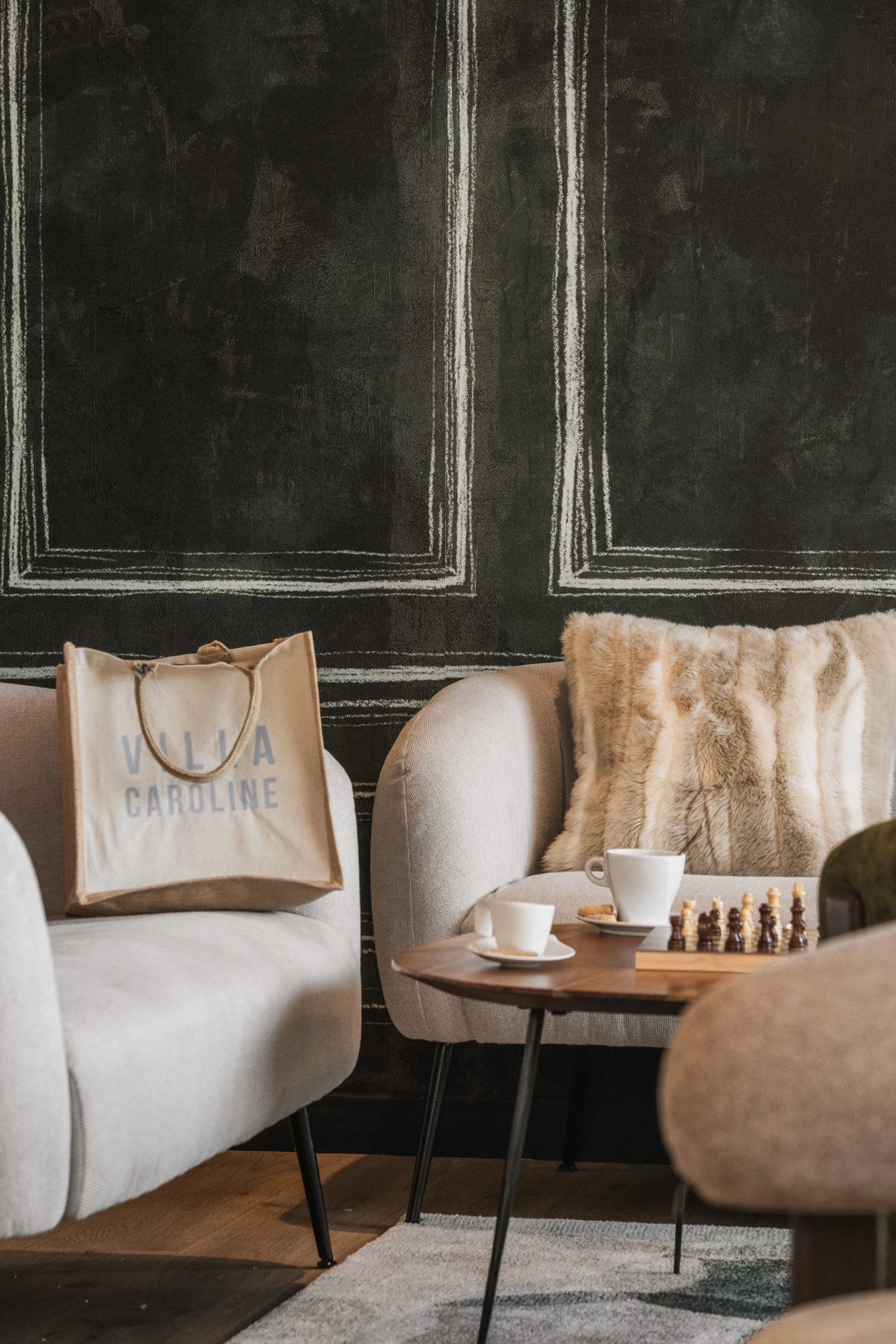 Living room with beige sofas, a wooden coffee table with cups and a chessboard, a fur throw pillow, a patterned rug, and a dark wall with white-lined borders.