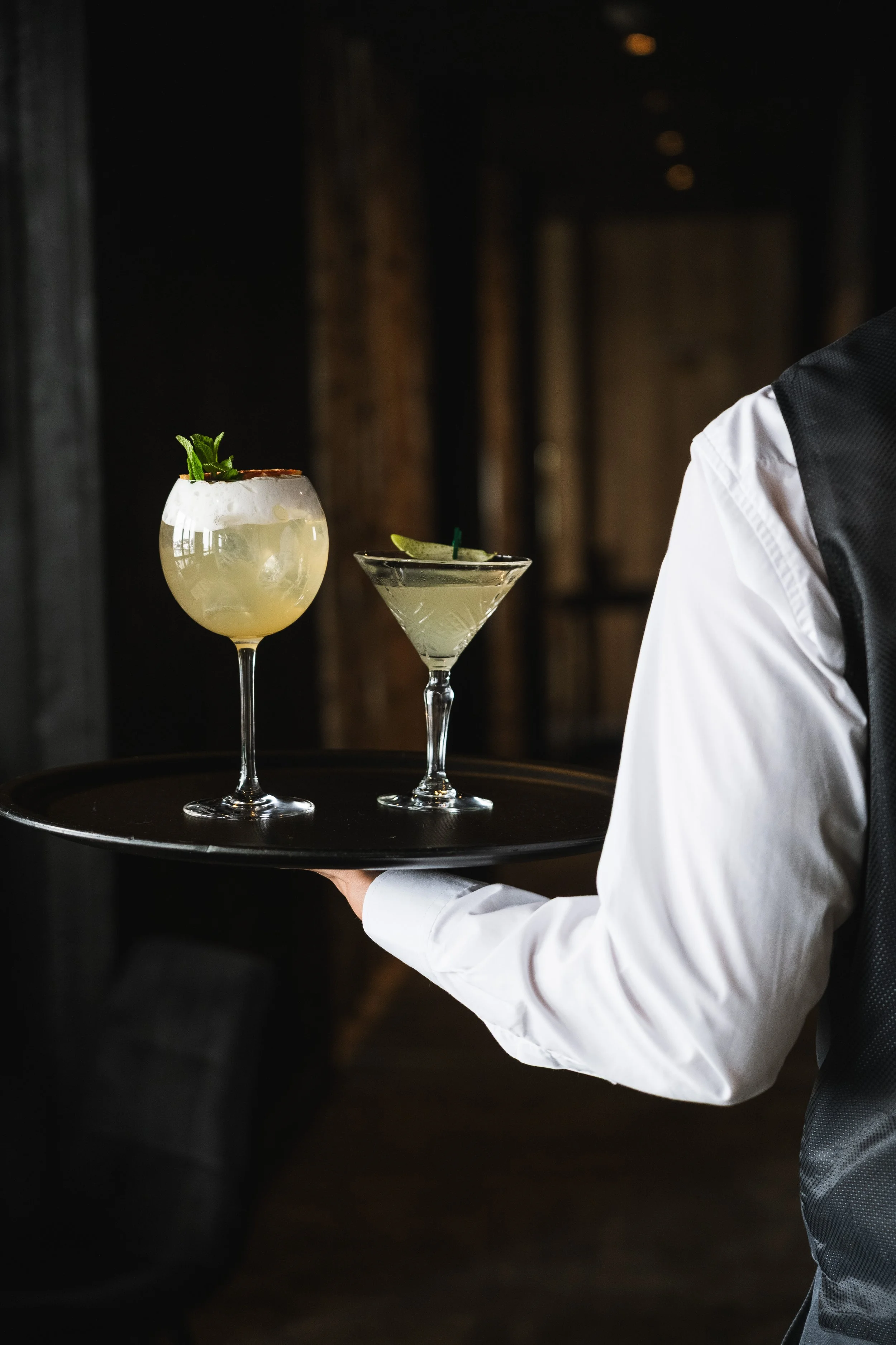 A waiter in a white shirt and dark vest holding a black tray with two cocktails, one garnished with mint and the other with a lime wedge, in a dimly lit bar or restaurant.