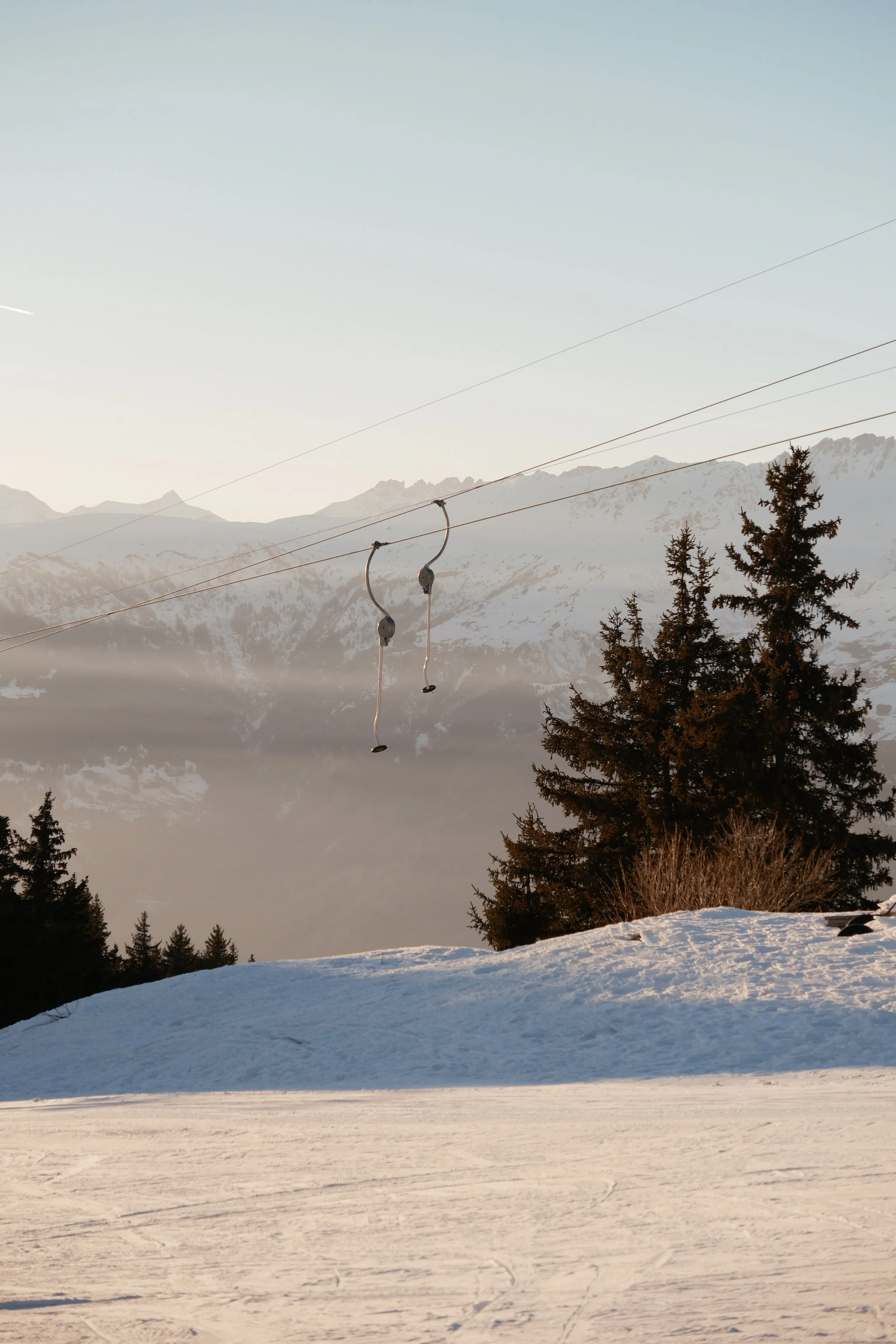 Snow-covered ground with scattered trees, mountains in the background, and ski lift cables overhead in a winter landscape.