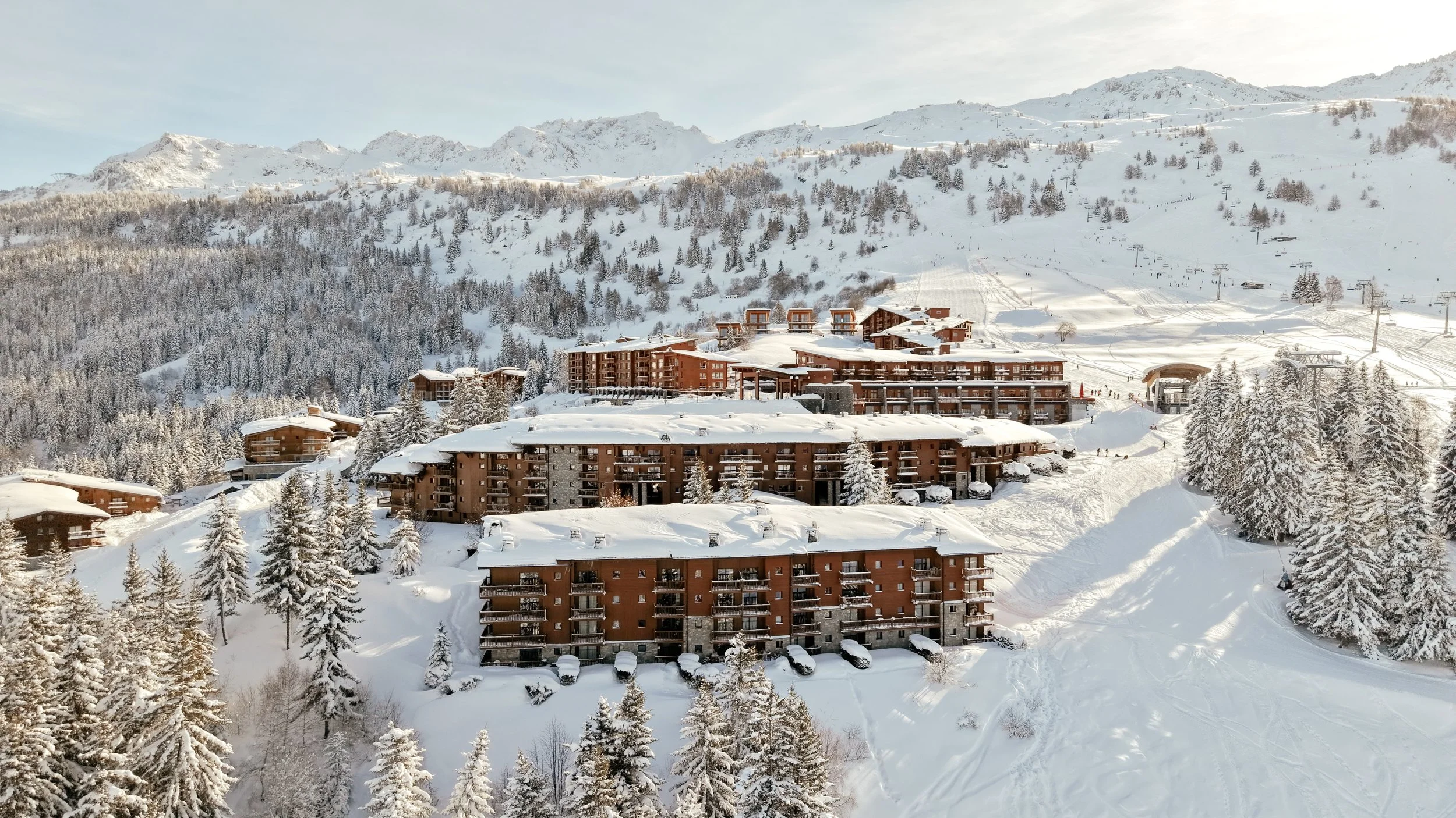 Snow-covered ski resort with chalet-style buildings surrounded by pine trees and snow-covered mountains in the background.