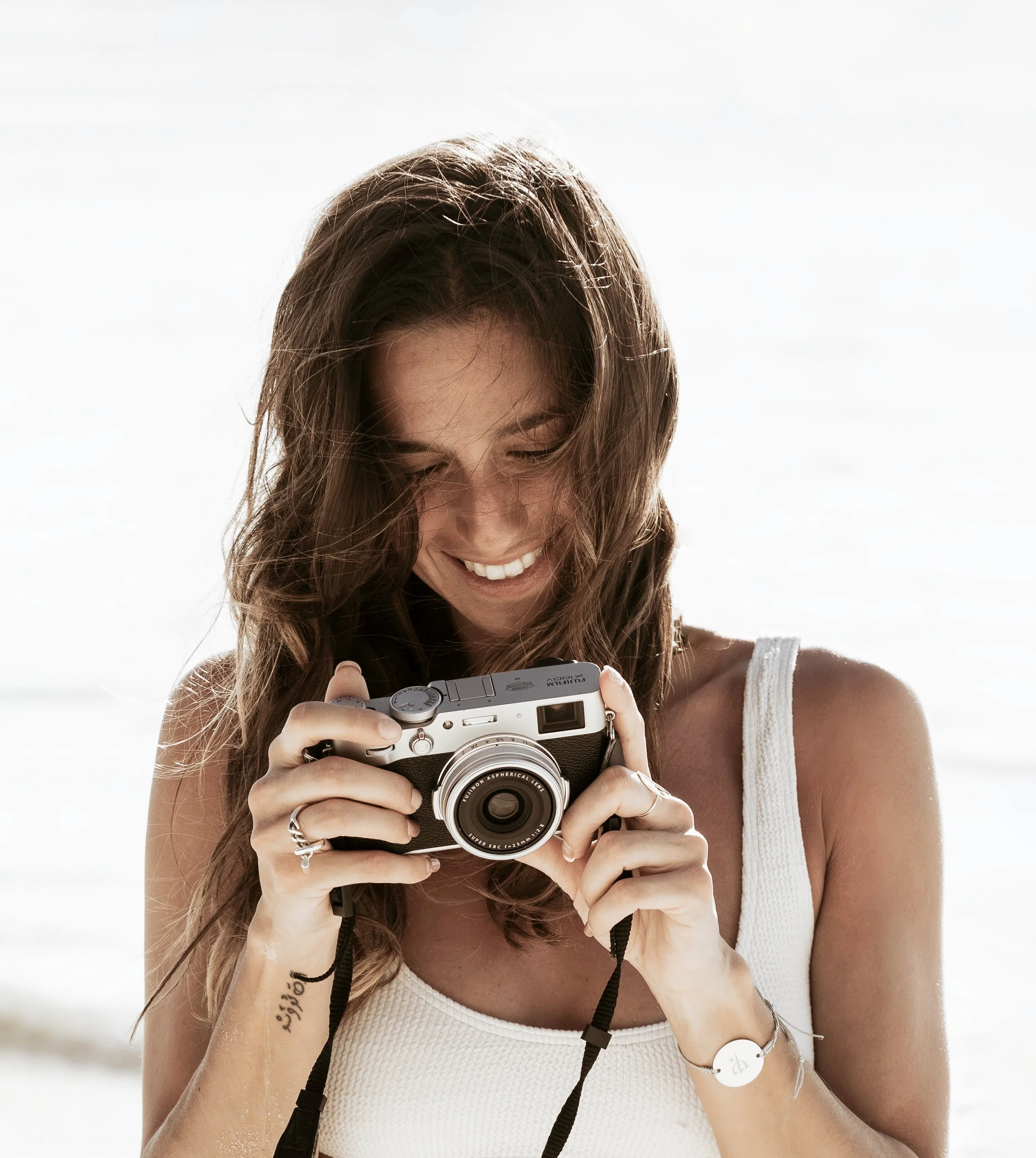 Young woman with long brown hair smiling while looking at a camera in her hands, wearing a white sleeveless top, outdoors with sky in the background.