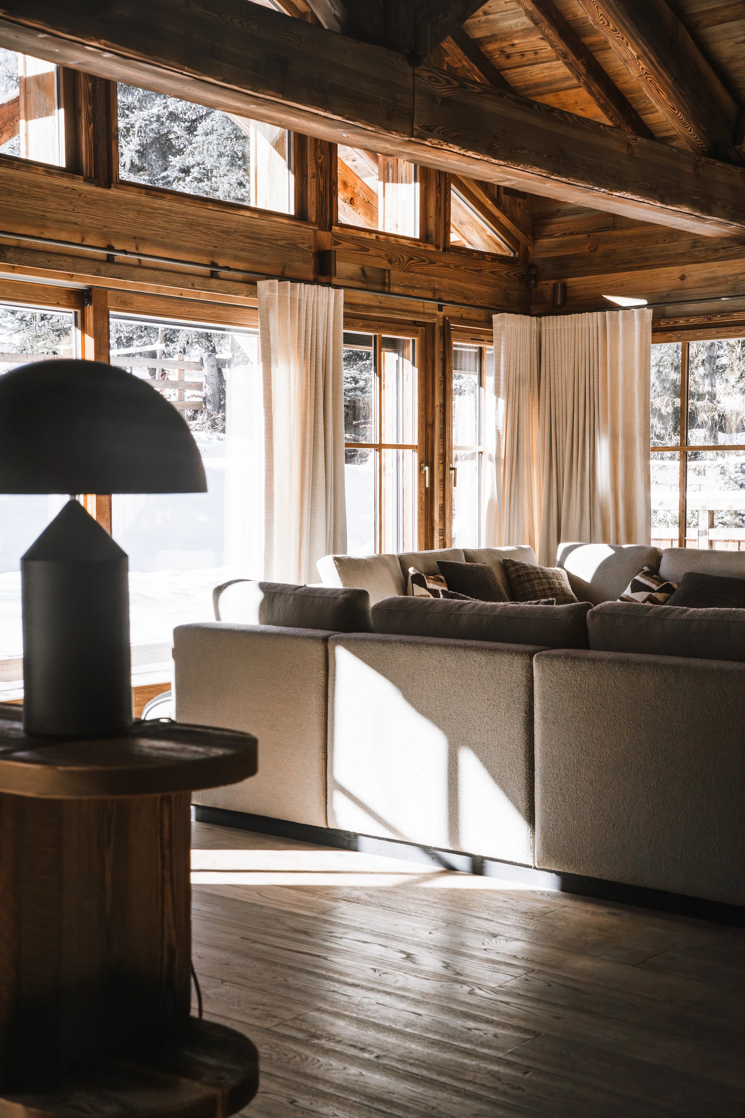 Living room with large windows overlooking a snowy landscape, wooden walls, ceiling, and floors, and beige sofas with patterned cushions.