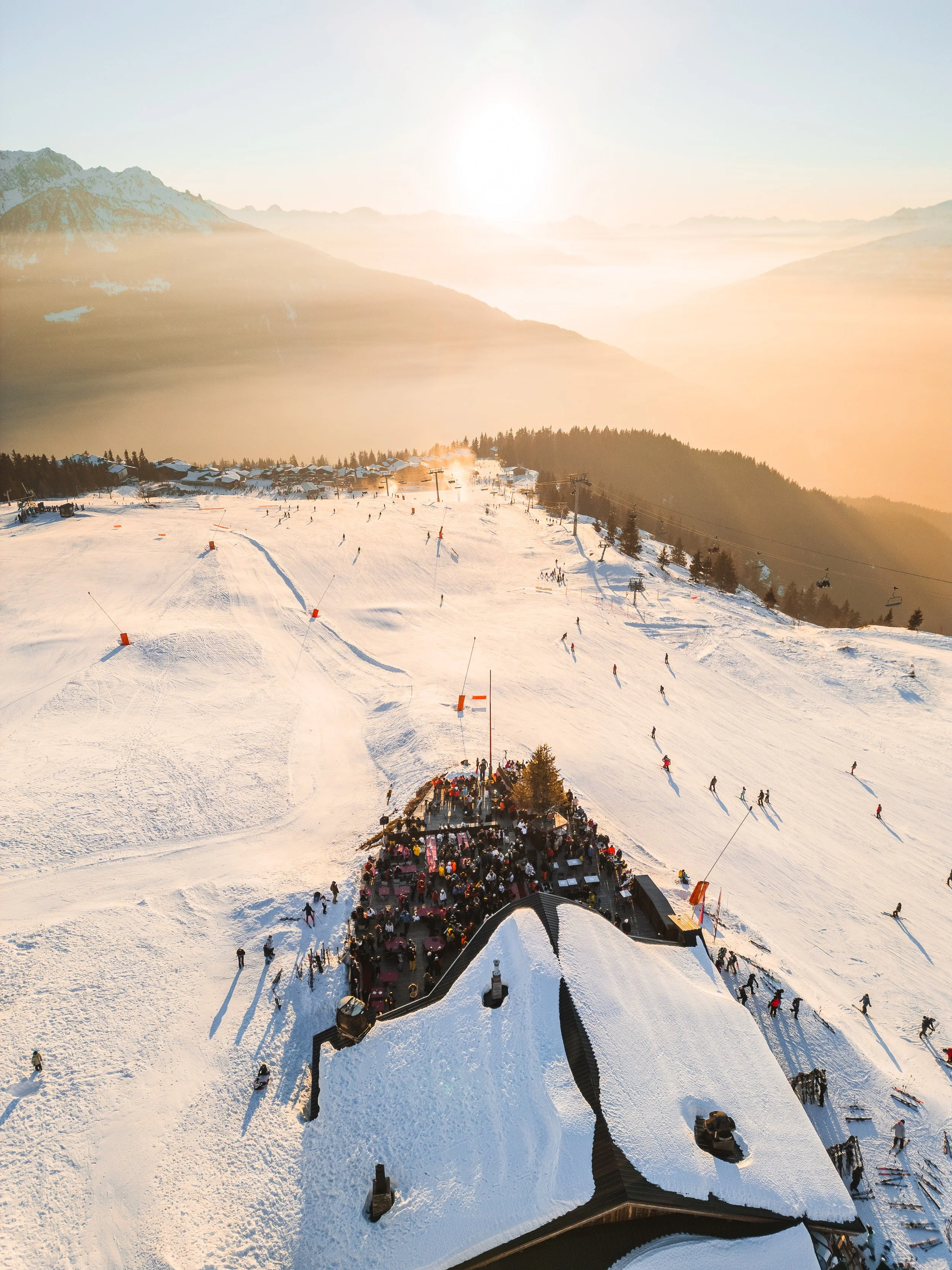Sunset over a snowy ski resort with skiers, snowboarders, ski lifts, and a large crowd gathered near a building with a black roof.