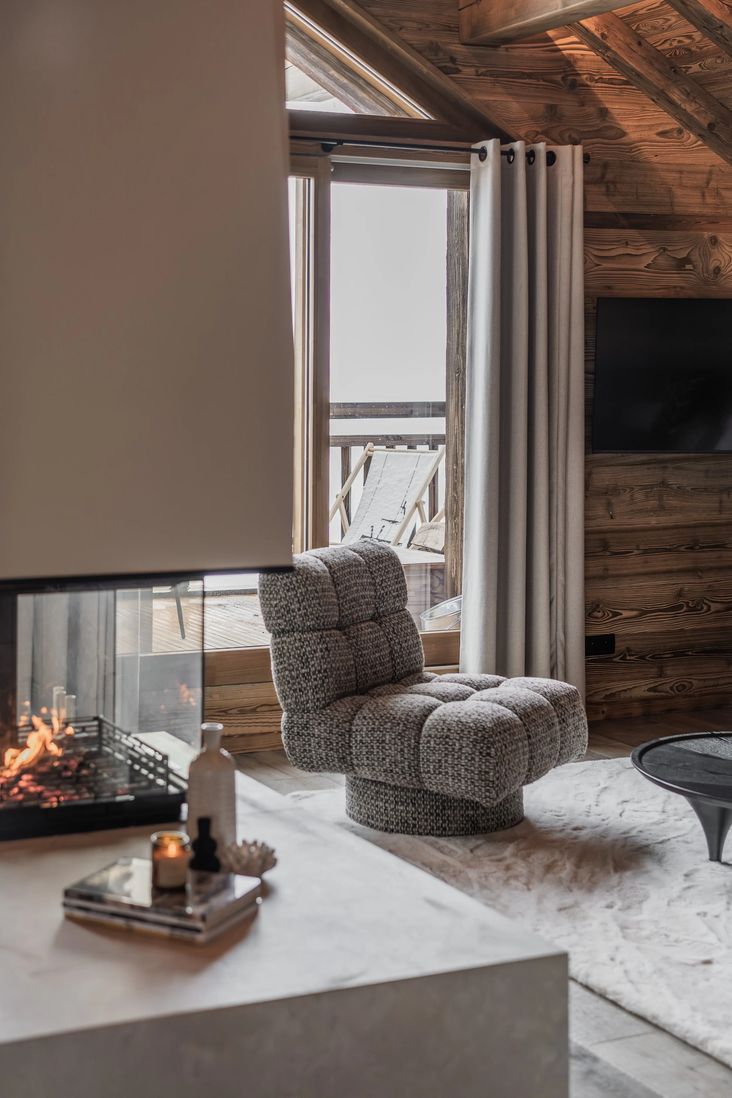 Living room with a cozy armchair, a fireplace, and a sliding glass door leading to a balcony with a lounge chair outside.