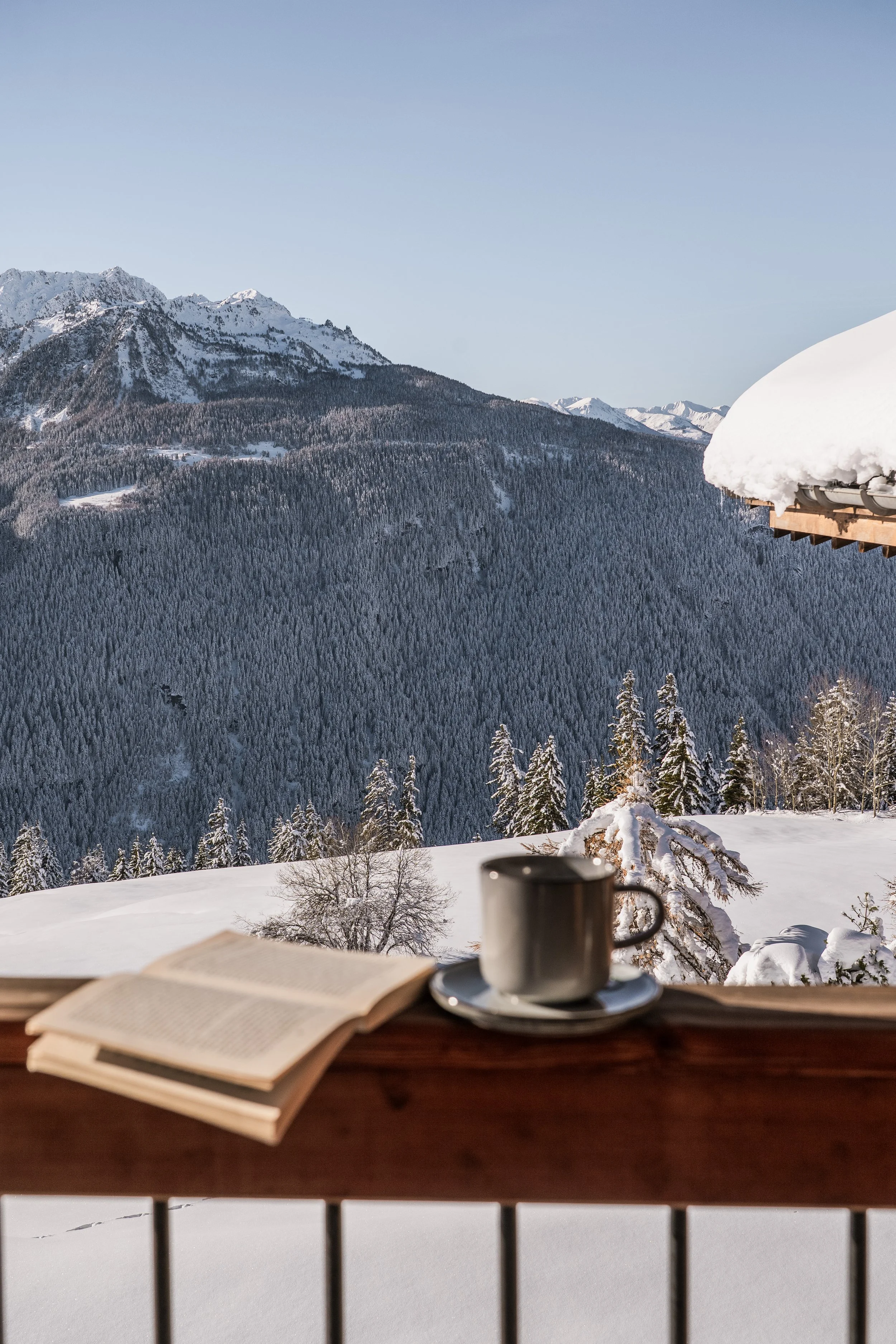 Snow-covered mountain landscape with trees, viewed from a balcony. In the foreground, a table holds an open book, a mug, and a saucer.