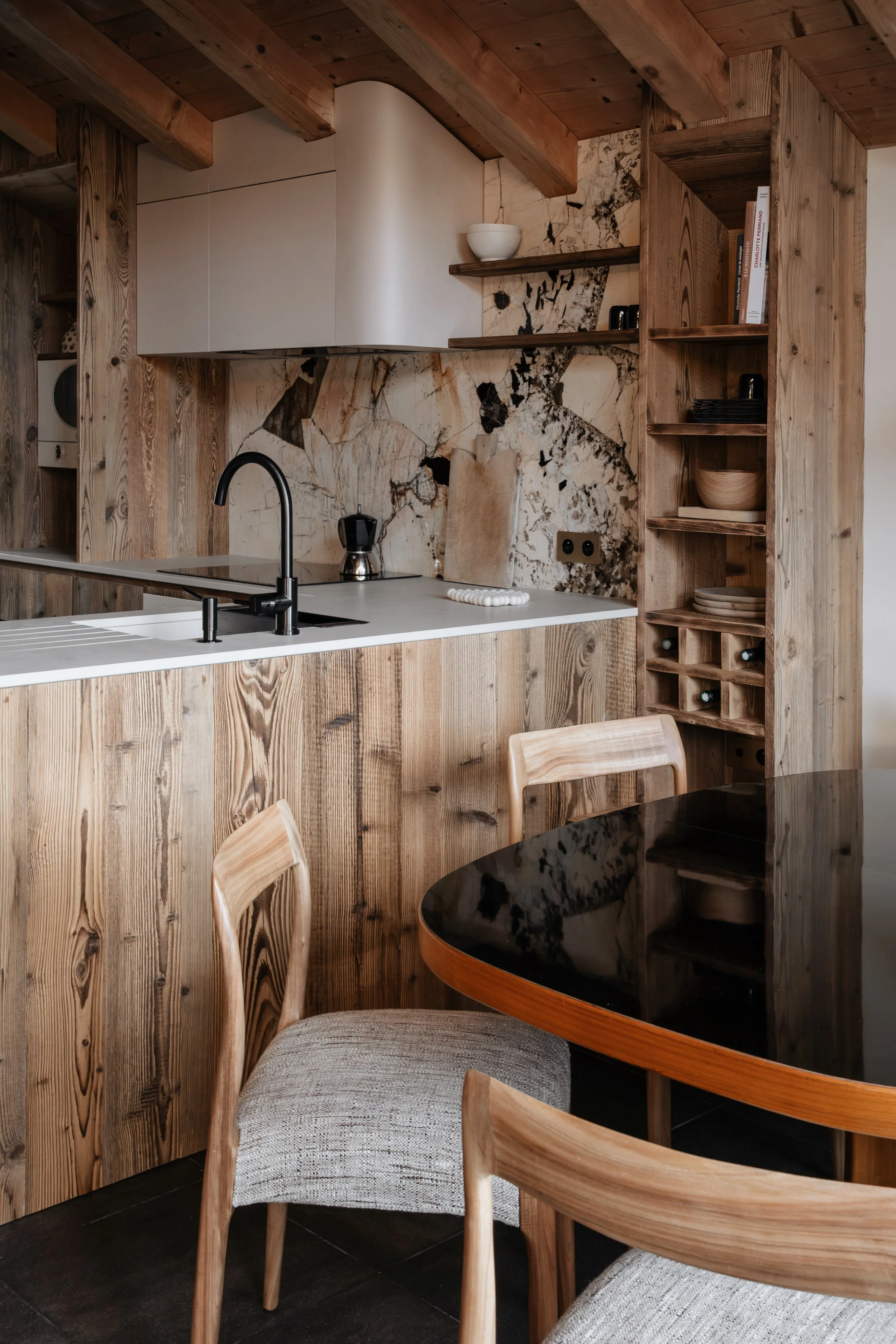 A kitchen with wooden cabinets, a marble backsplash, open shelves with books and bowls, a black sink faucet, a chopping board, and a coffee maker. Part of a dining table with wooden chairs is visible.
