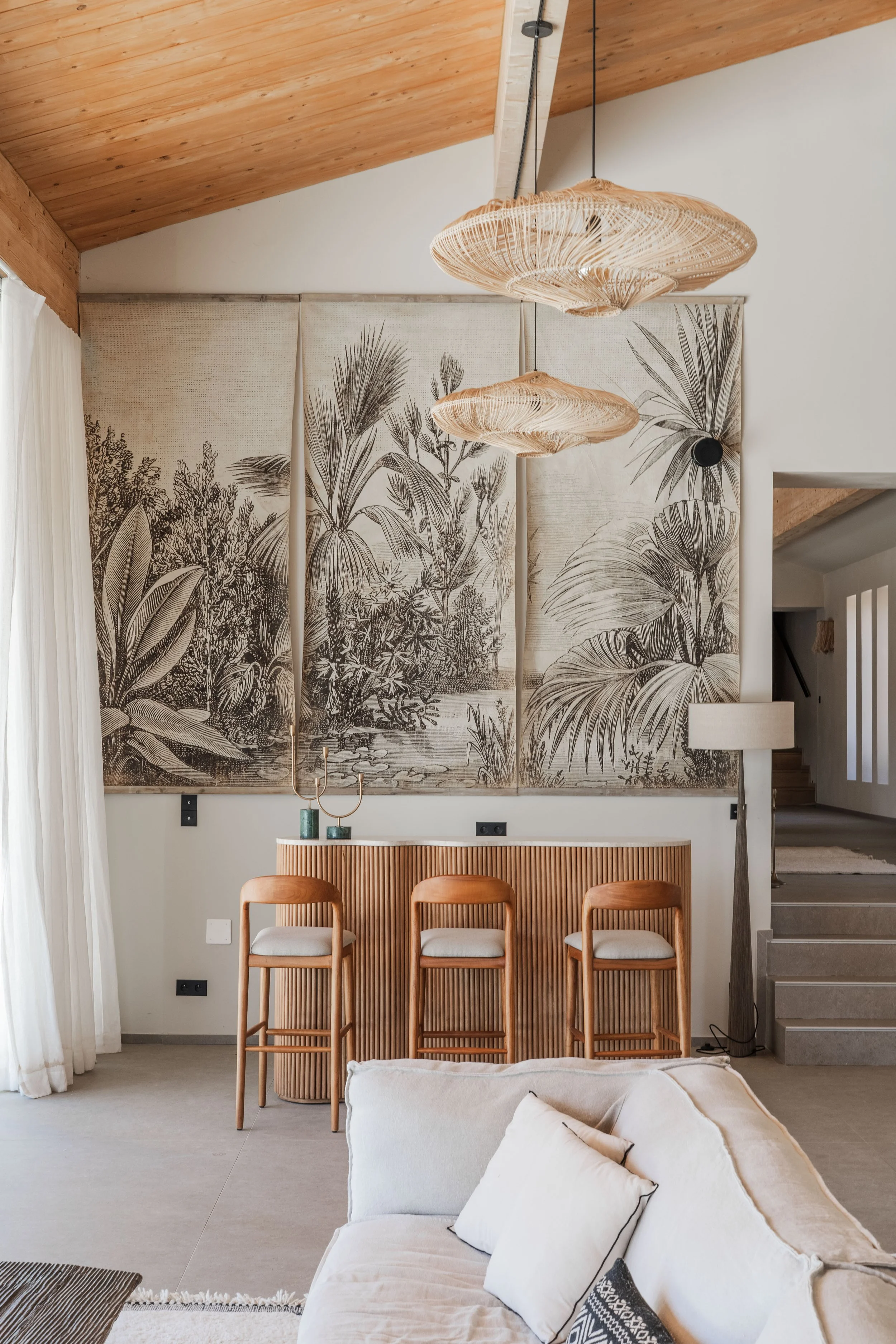 Living room with white sofas, floral black-and-white curtains, wooden chairs with white cushions, bamboo pendant lights, and botanical-themed wall art.