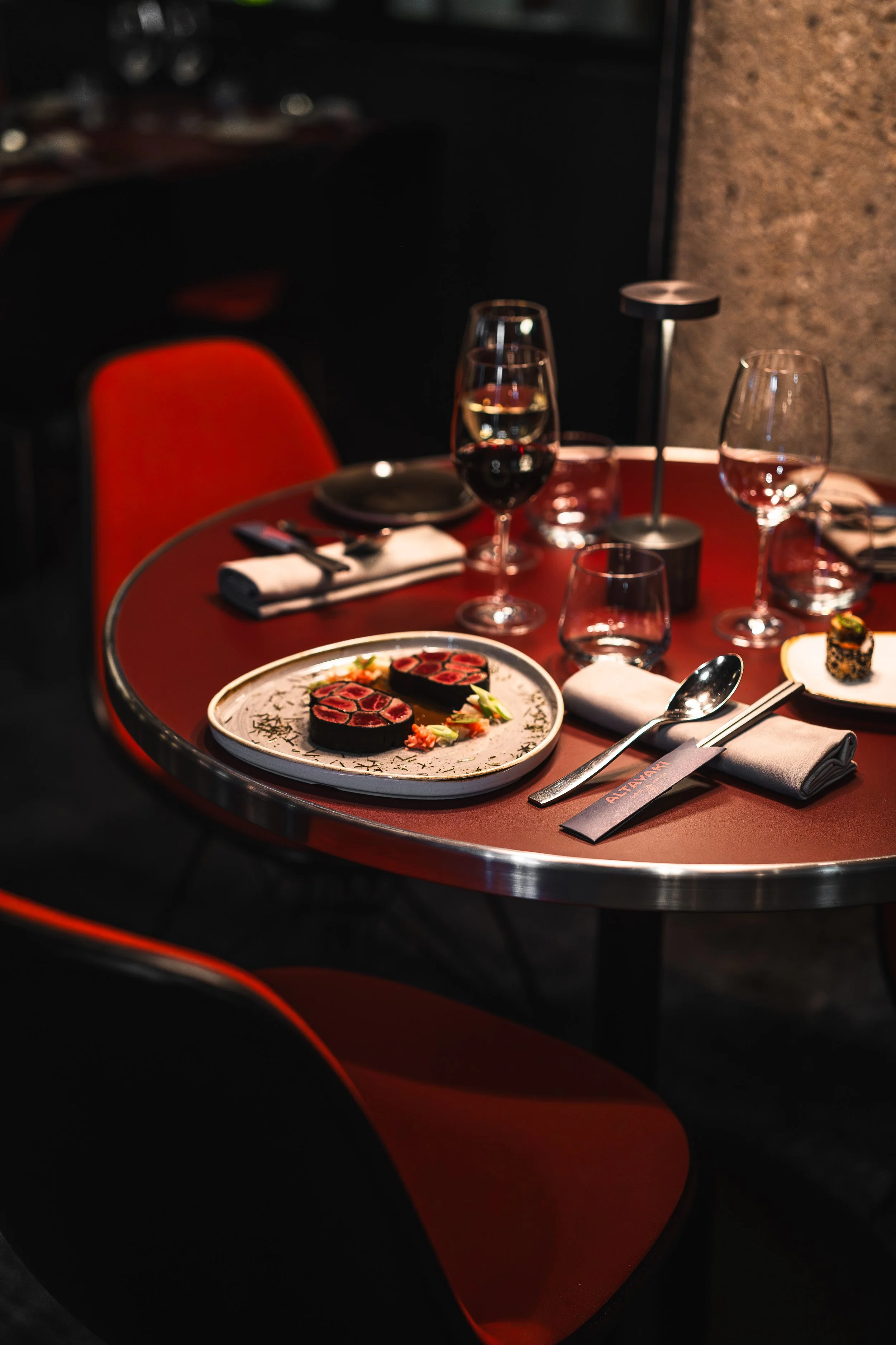 A dinner table with plates, glasses of wine, and chopsticks, set in a dimly lit restaurant with a modern interior featuring a red chair and a stone wall.