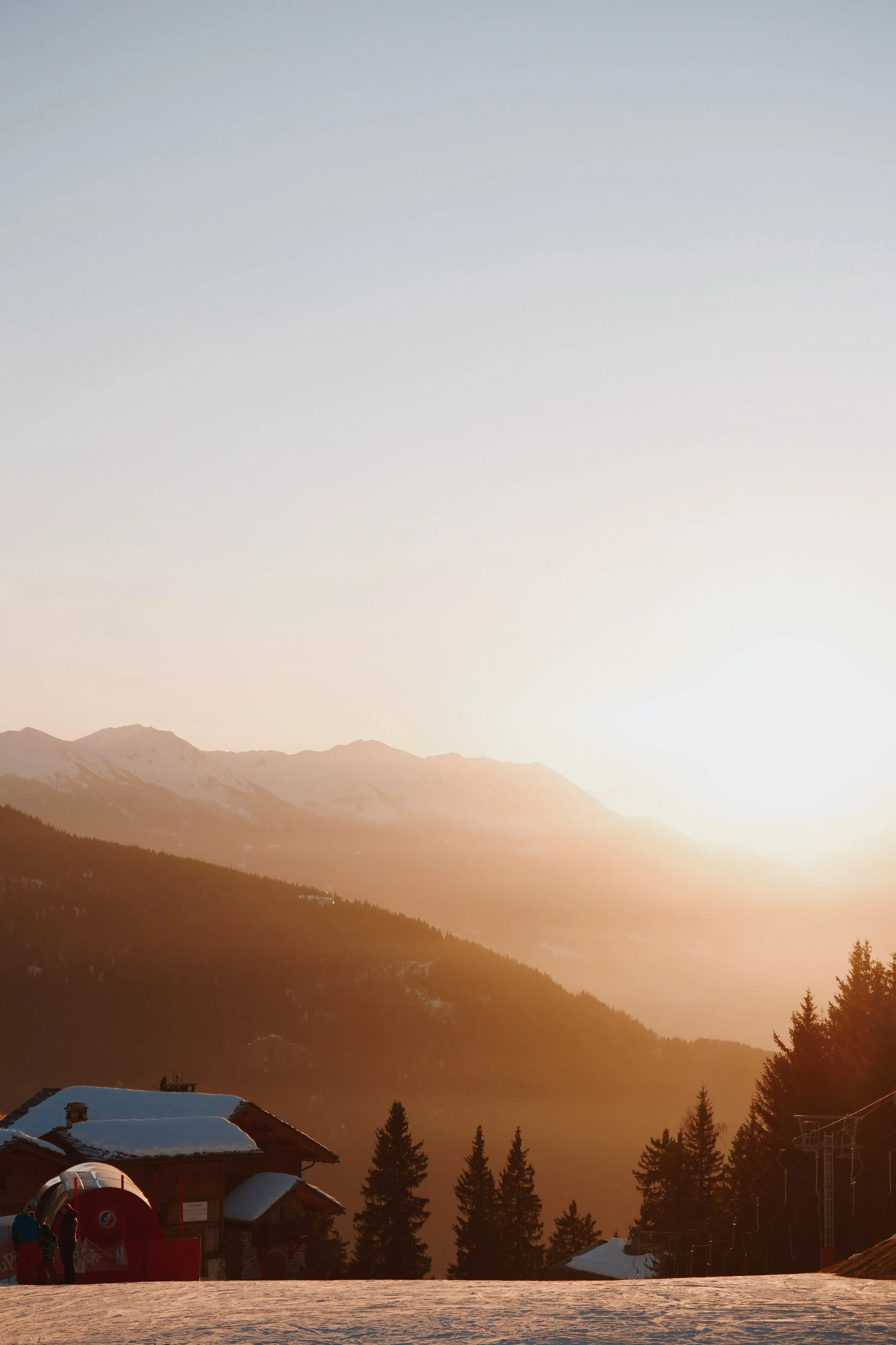 Snow-covered rooftops and ski equipment in front of a mountainous landscape at sunset, with trees and a ski slope in the foreground.