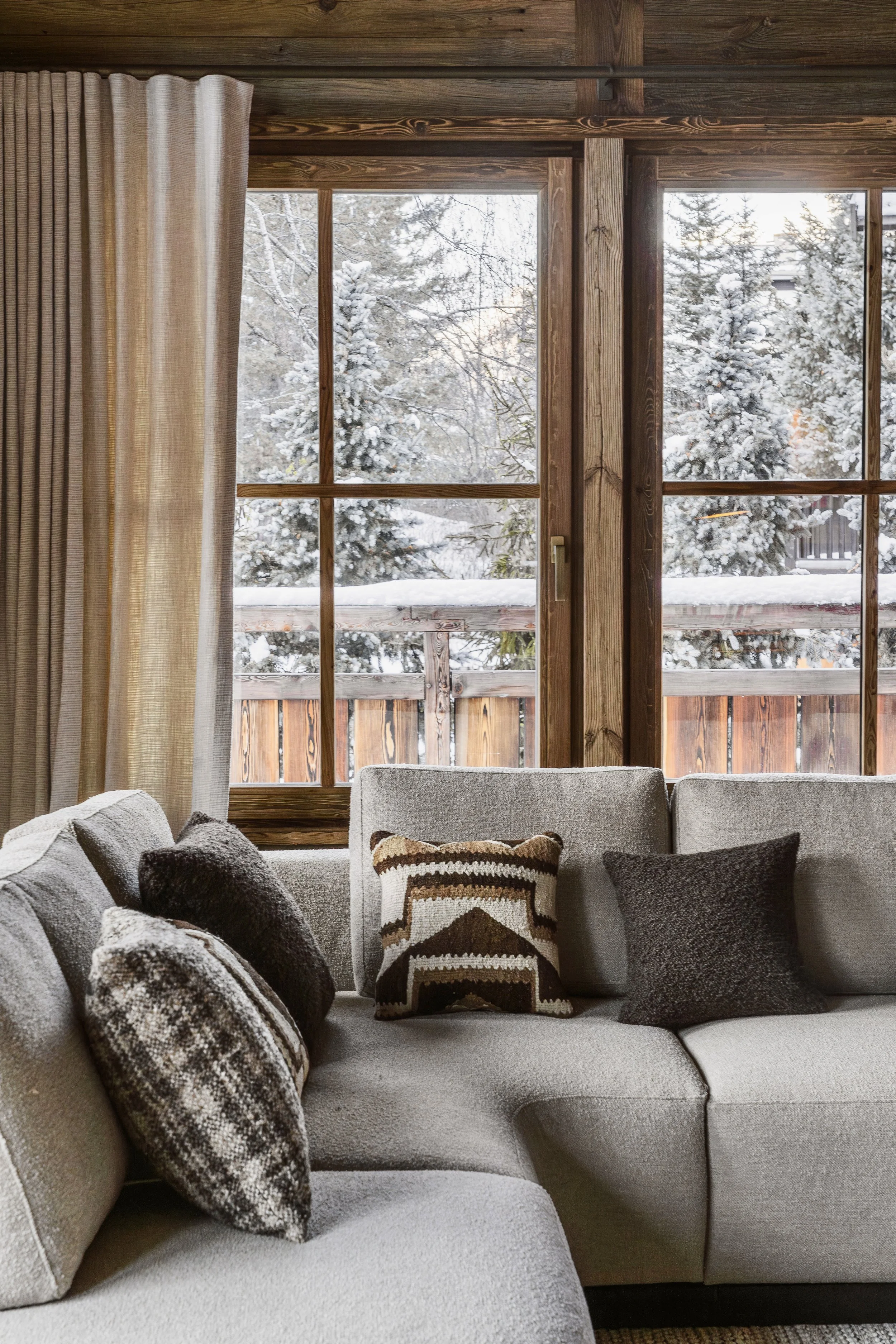 Living room with beige sectional sofa with decorative pillows, facing large window with wooden frame and beige curtains, overlooking snow-covered trees outside.