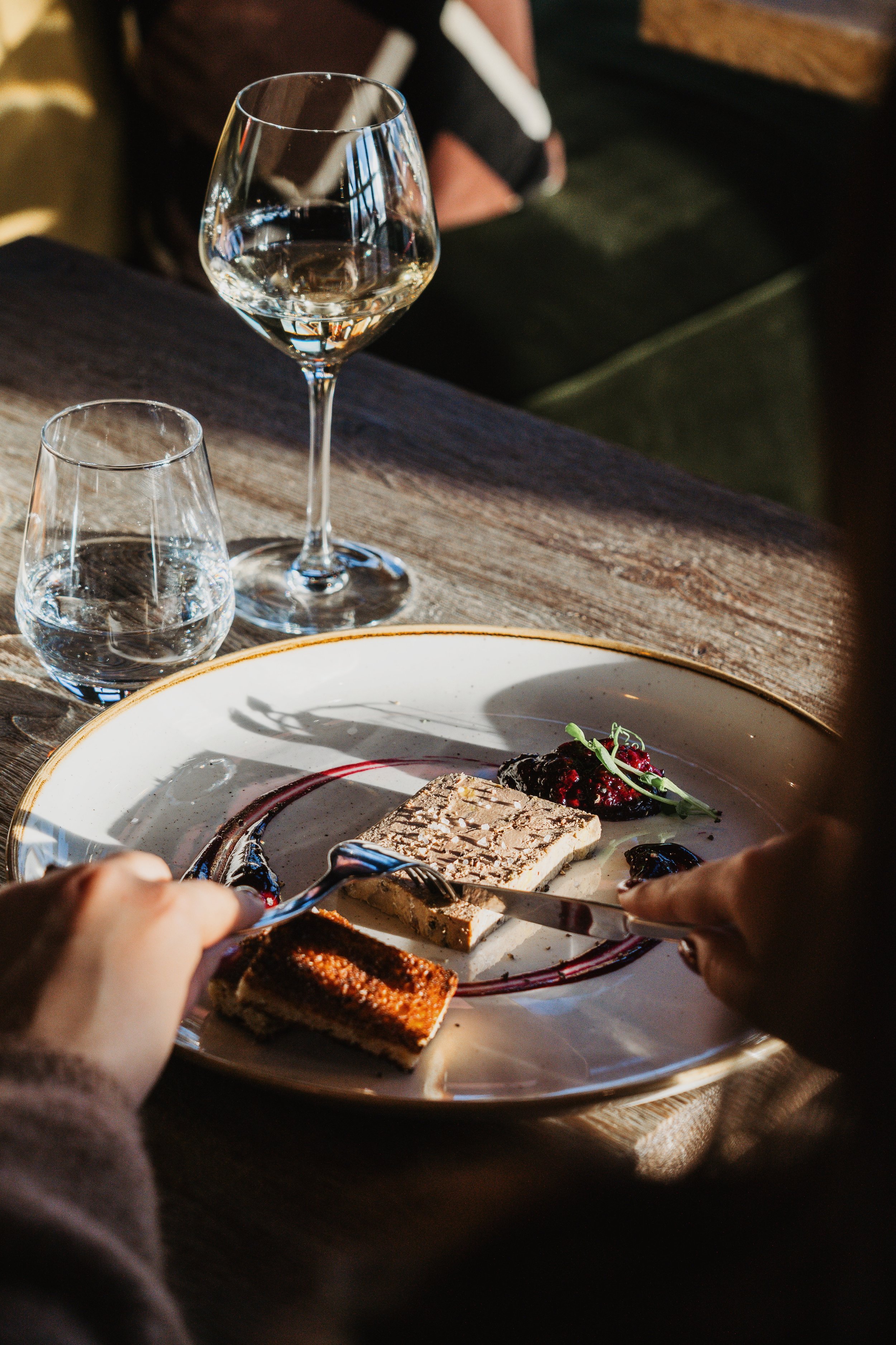 A dining scene with a plate of assorted desserts, including a layered cake, berry compote, and chocolate treat, on a white plate with a decorative border. There are two hands holding utensils, cutting and serving the desserts. In the background, ther