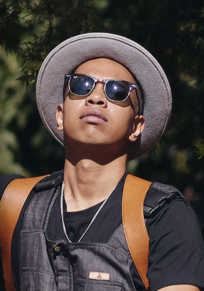 Young man wearing sunglasses, a wide-brimmed hat, a black backpack, and a black T-shirt, outdoors with trees in the background.
