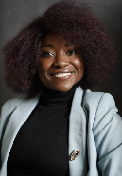 A smiling woman with natural, curly hair wearing a black turtleneck and a light-colored blazer posing against a dark background.