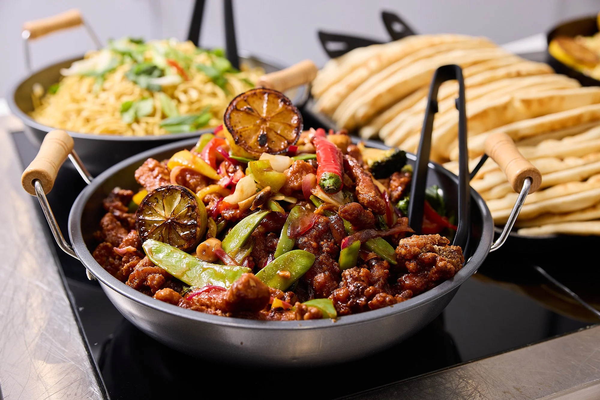 A serving tray with three black bowls containing Chinese or Asian-inspired dishes: one with stir-fried vegetables and chicken, another with fried noodles, and the third with crispy egg rolls.