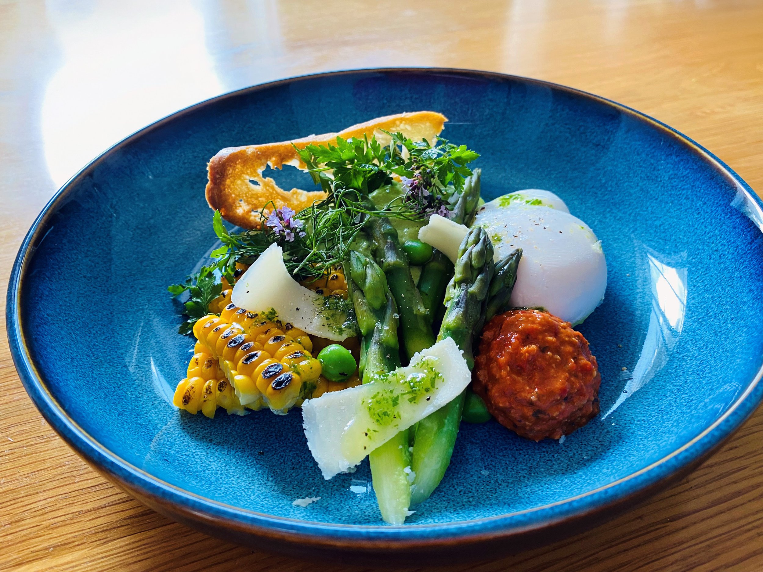 A blue bowl with a gourmet dish containing grilled corn, poached eggs, asparagus, fresh herbs, and a meatball, placed on a wooden surface.