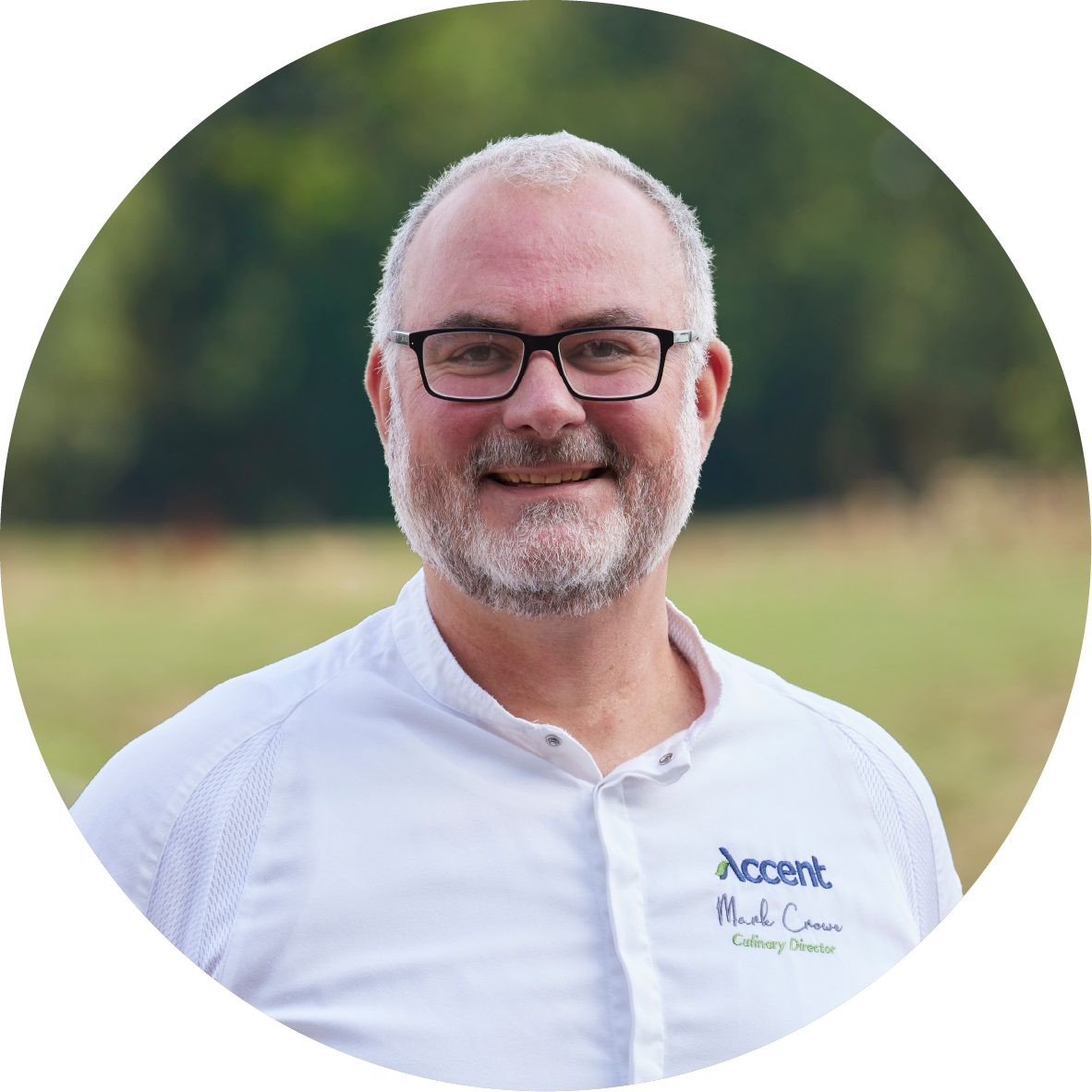 A smiling middle-aged man with glasses, gray hair, and a beard, wearing a white shirt with the logo 'Accent' and the text 'Culinary Director', standing outdoors with blurred green trees and grass in the background.