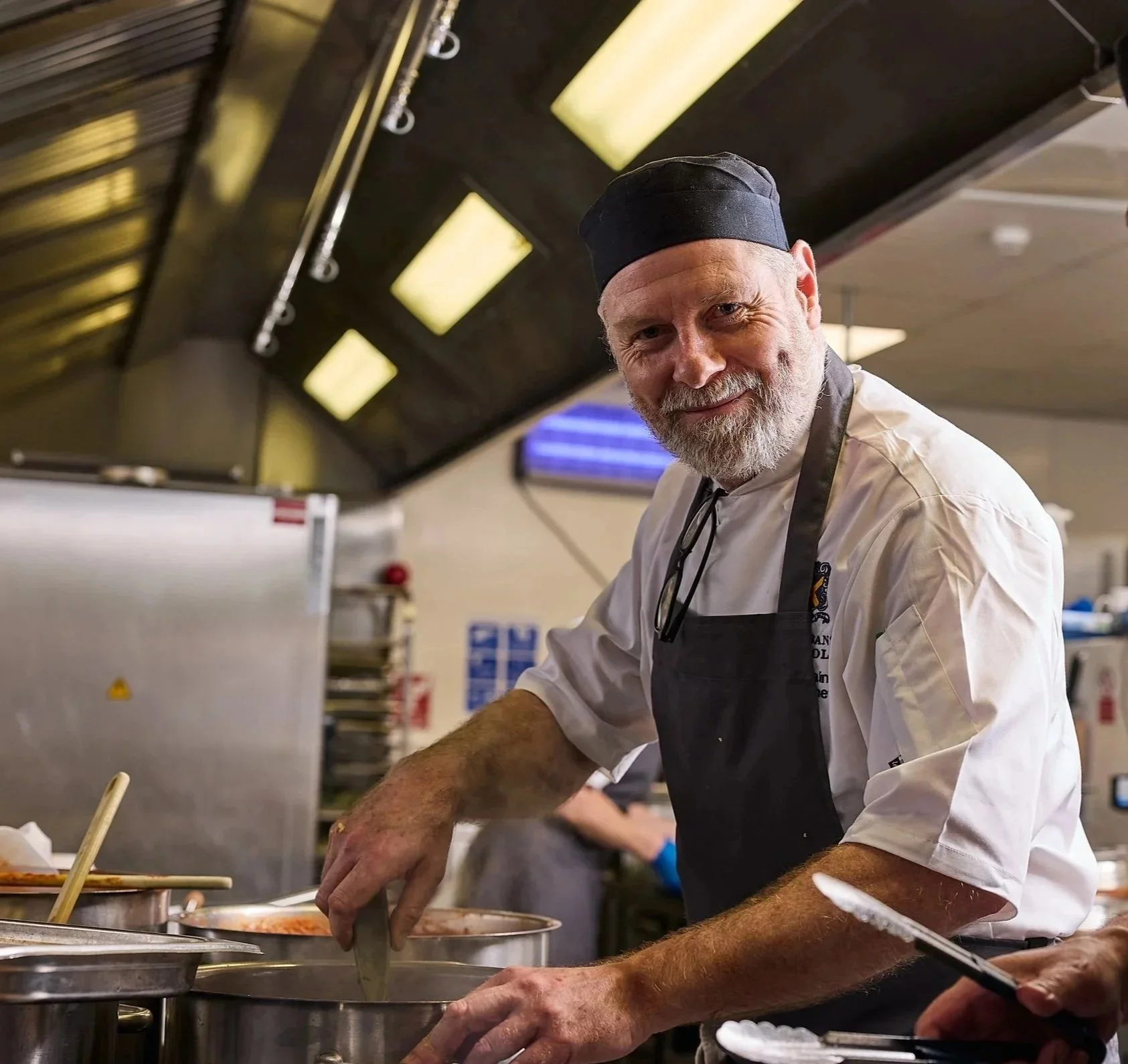 A smiling chef with a gray beard and black chef's hat, wearing a white chef's coat and black apron, preparing food in a commercial kitchen.