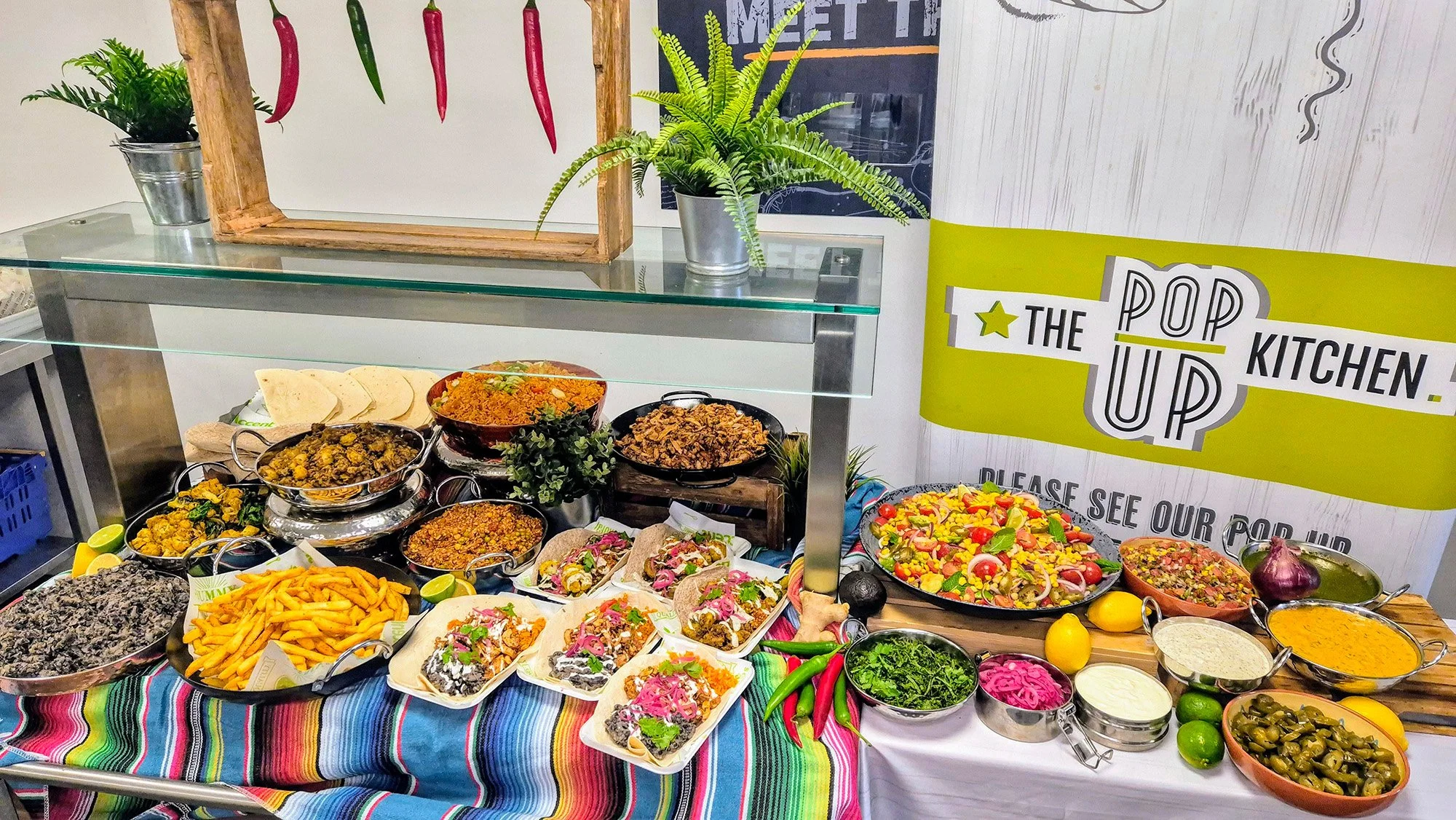 Display of various Mexican dishes and condiments on a table with a colorful striped tablecloth, including rice, tortillas, salads, fried foods, and beans, with chili peppers and plants in the background.