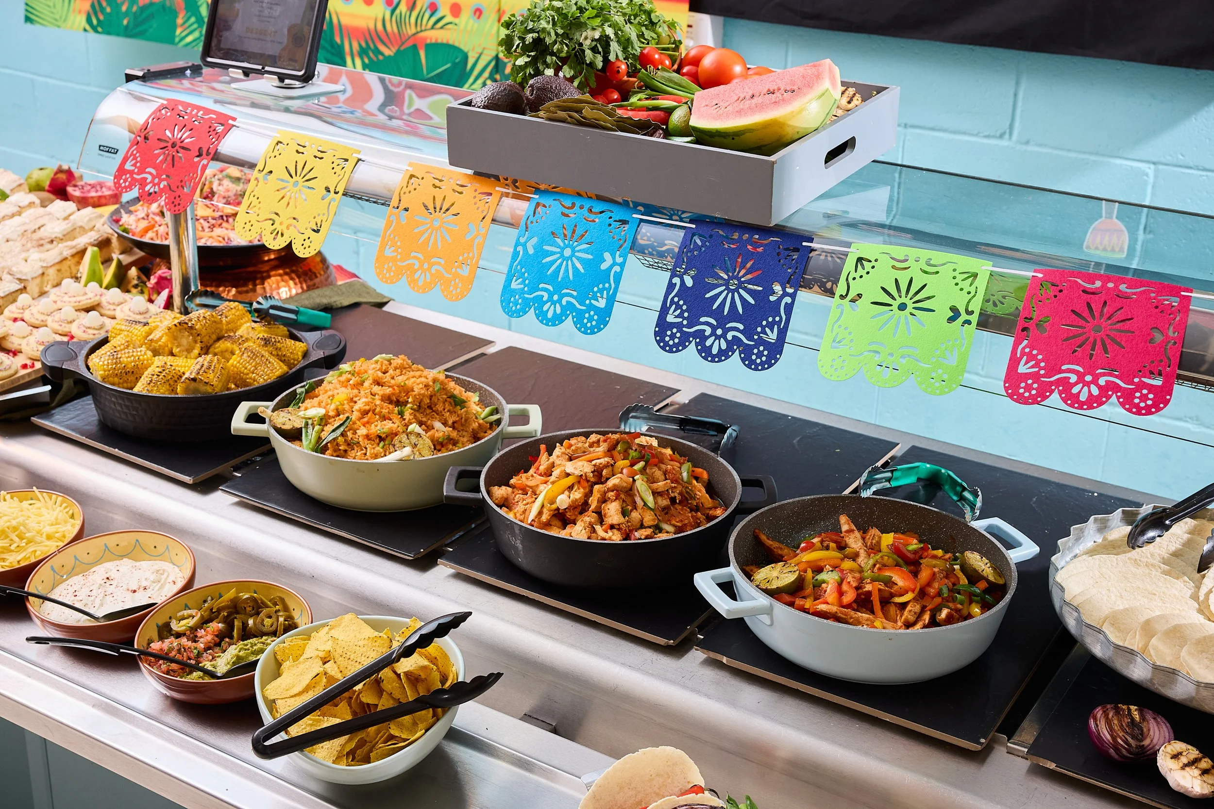 A buffet of Mexican food including tacos, rice, vegetables, and chips, with colorful paper decorations hanging above.