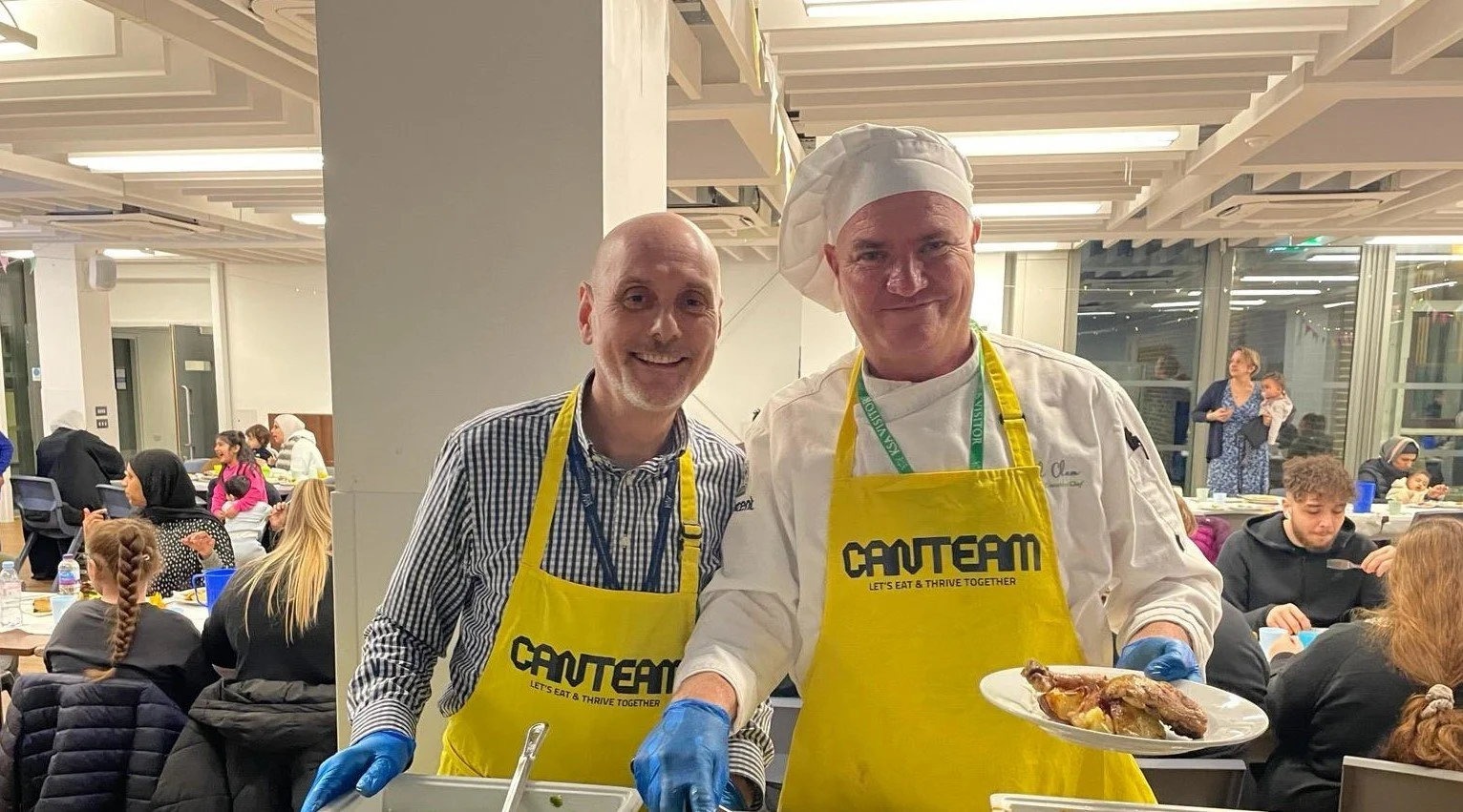 Two smiling men in yellow aprons and chef attire serving food at a communal event, with others seated at tables in a well-lit indoor space.