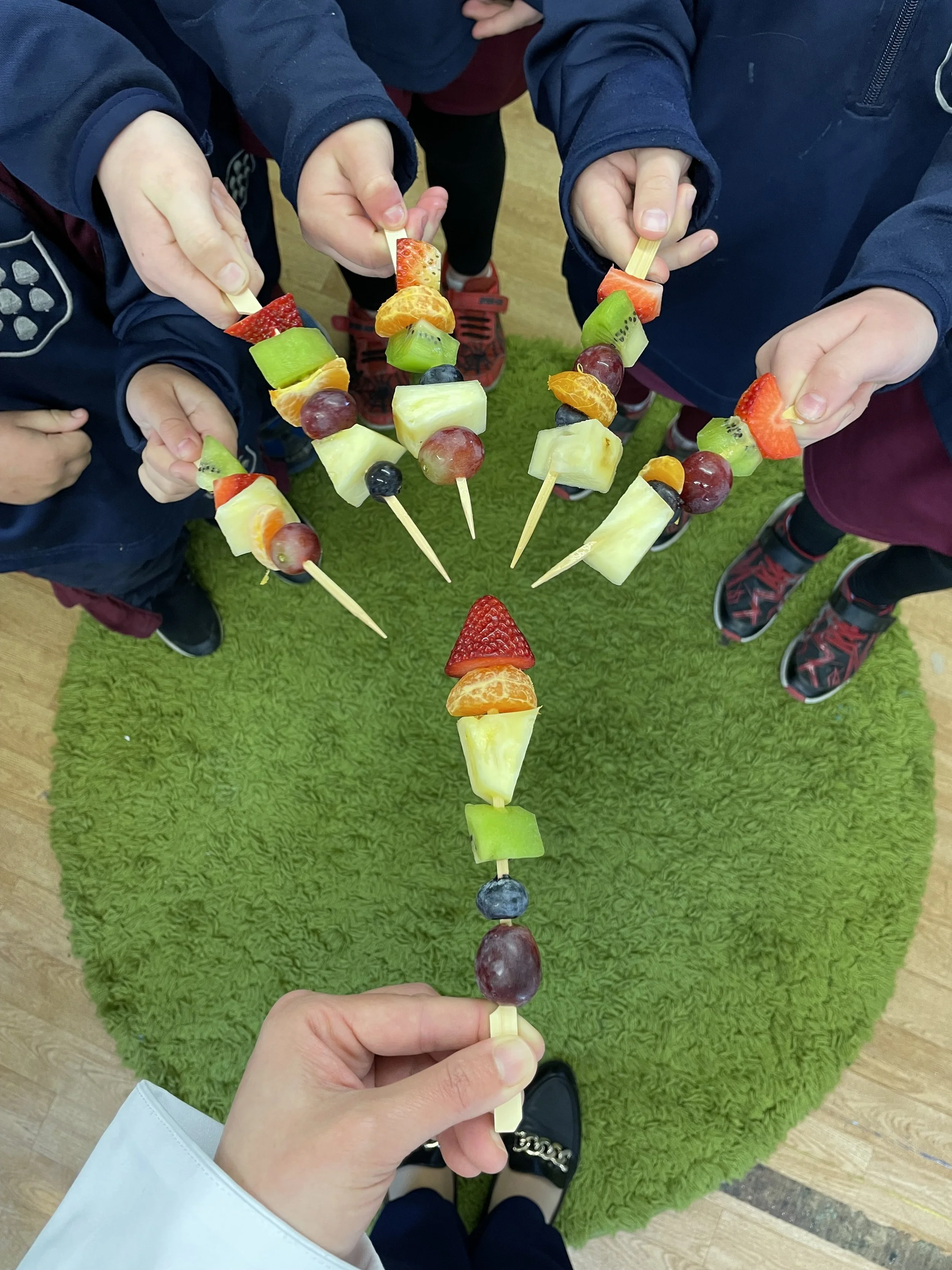 Close-up of a hand holding a fruit skewer with various fruits including blueberries, grapes, kiwi, orange, pineapple, strawberry, and watermelon, with several other people holding similar fruit skewers around a green carpet.