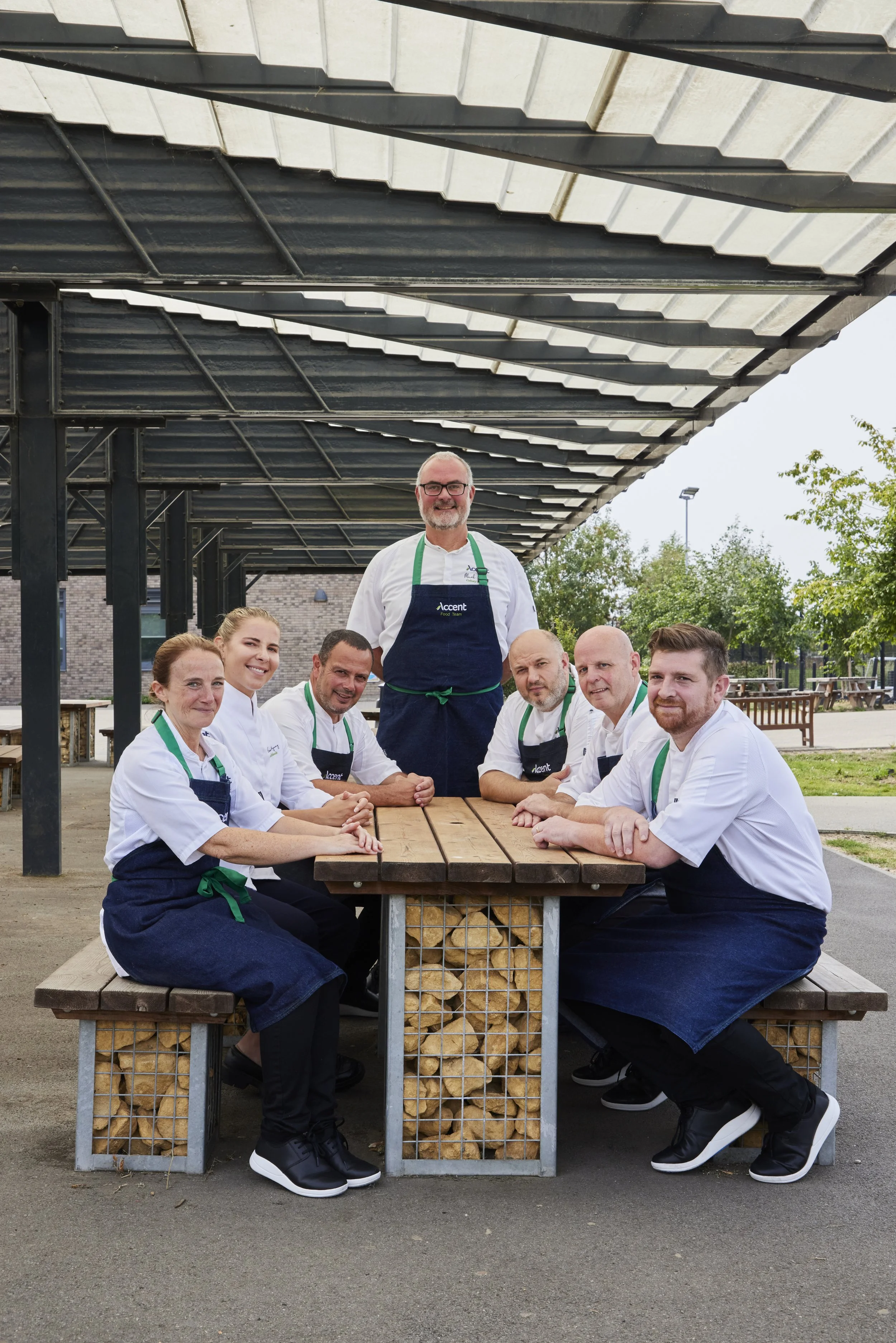 A group of eight people dressed as chefs sitting and standing around a wooden table outdoors, wearing white chef coats, black aprons, and black shoes. One person is standing behind them, all smiling. They are under a metal and glass outdoor canopy with a partly cloudy sky and trees in the background.