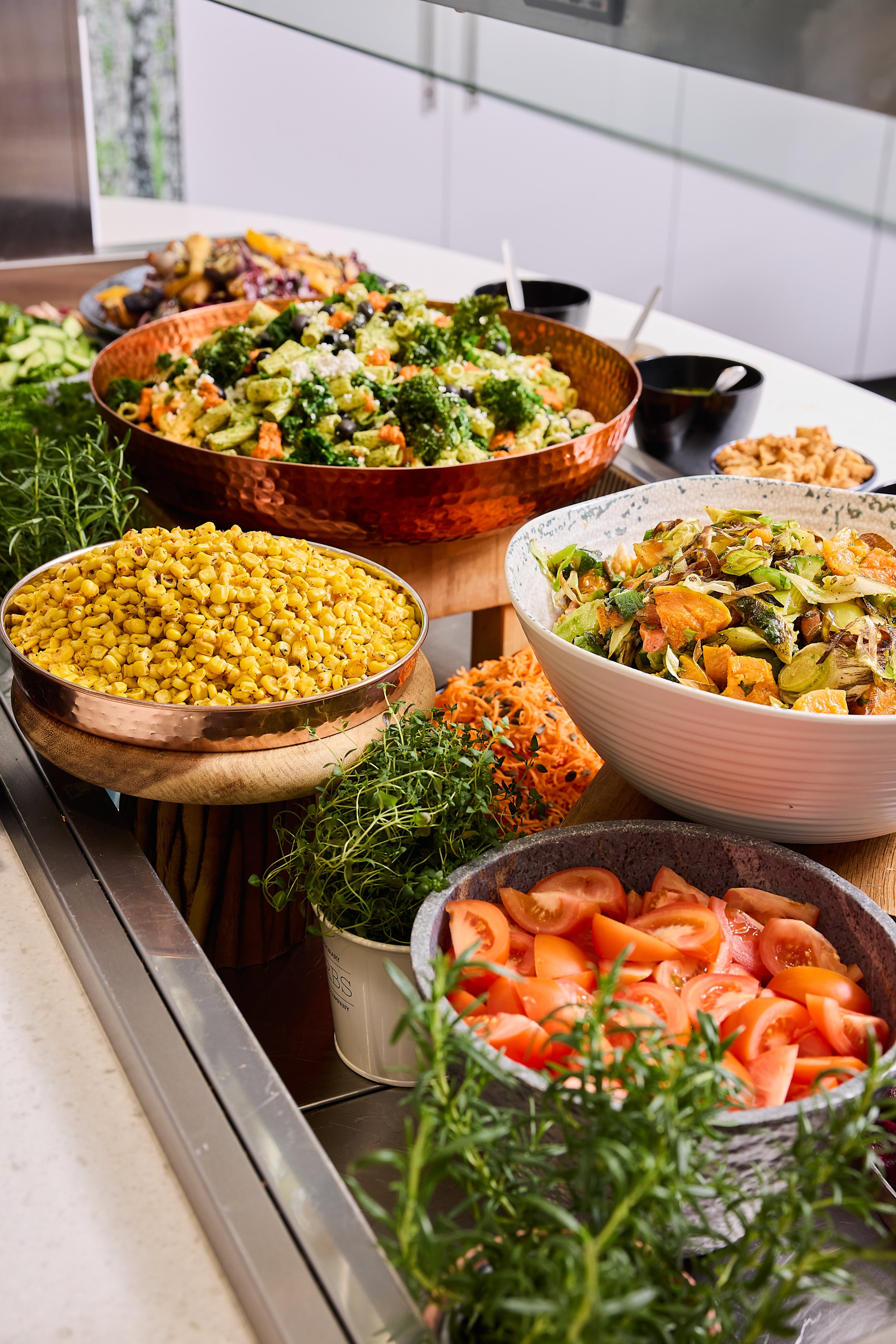 Assorted salads and vegetables on display, including chopped tomatoes, cooked corn, leafy greens, pasta salad with broccoli, and other mixed vegetables in bowls, set on a kitchen countertop.