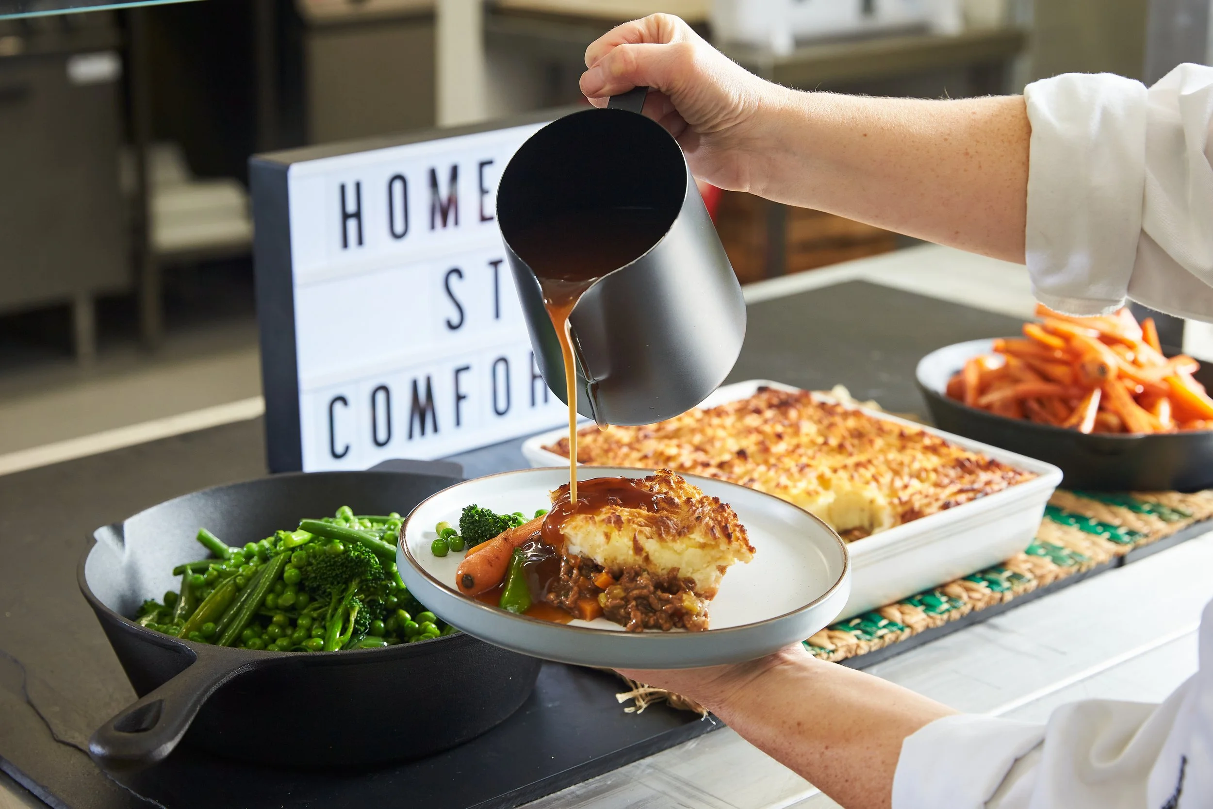 Person pouring gravy over a serving of mashed potatoes with mixed vegetables, with other baked dishes and a sign that reads 'Home Sweet Comfort' in the background.