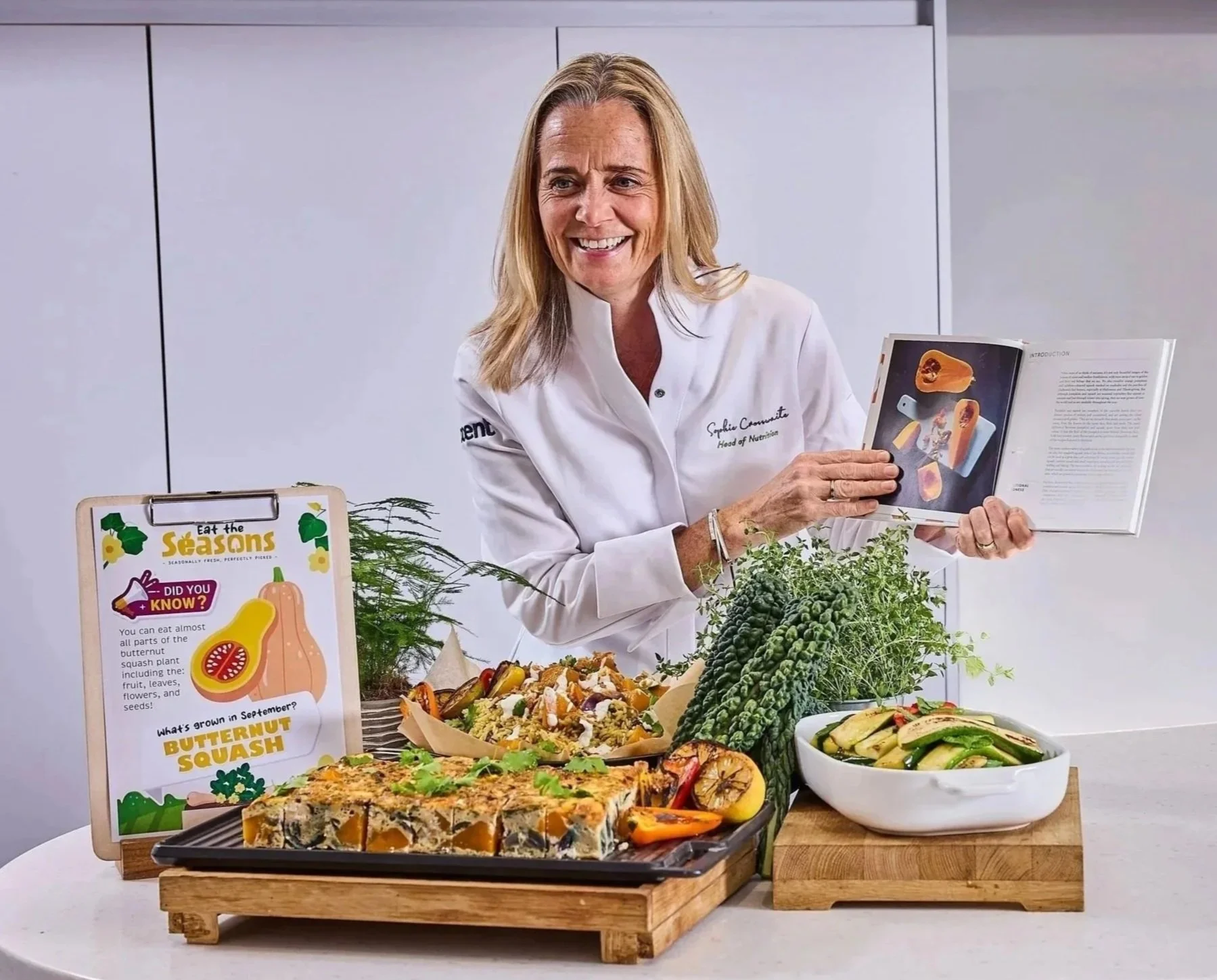 A woman wearing a white chef coat standing behind a table with various dishes and plants. She is holding a book open and smiling. The table has food items including a vegetable quiche, salad, and other healthy dishes, with a sign about seasonal squash and butternut squash.