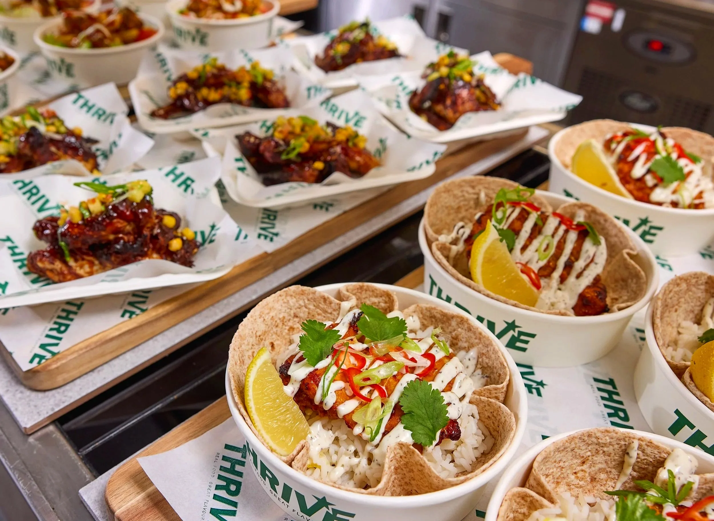 Close-up of multiple bowls of Mexican street food, including tacos with rice, garnished with lemon, cilantro, and sliced peppers, and cups of meat with sauce and toppings, arranged on a wooden tray.