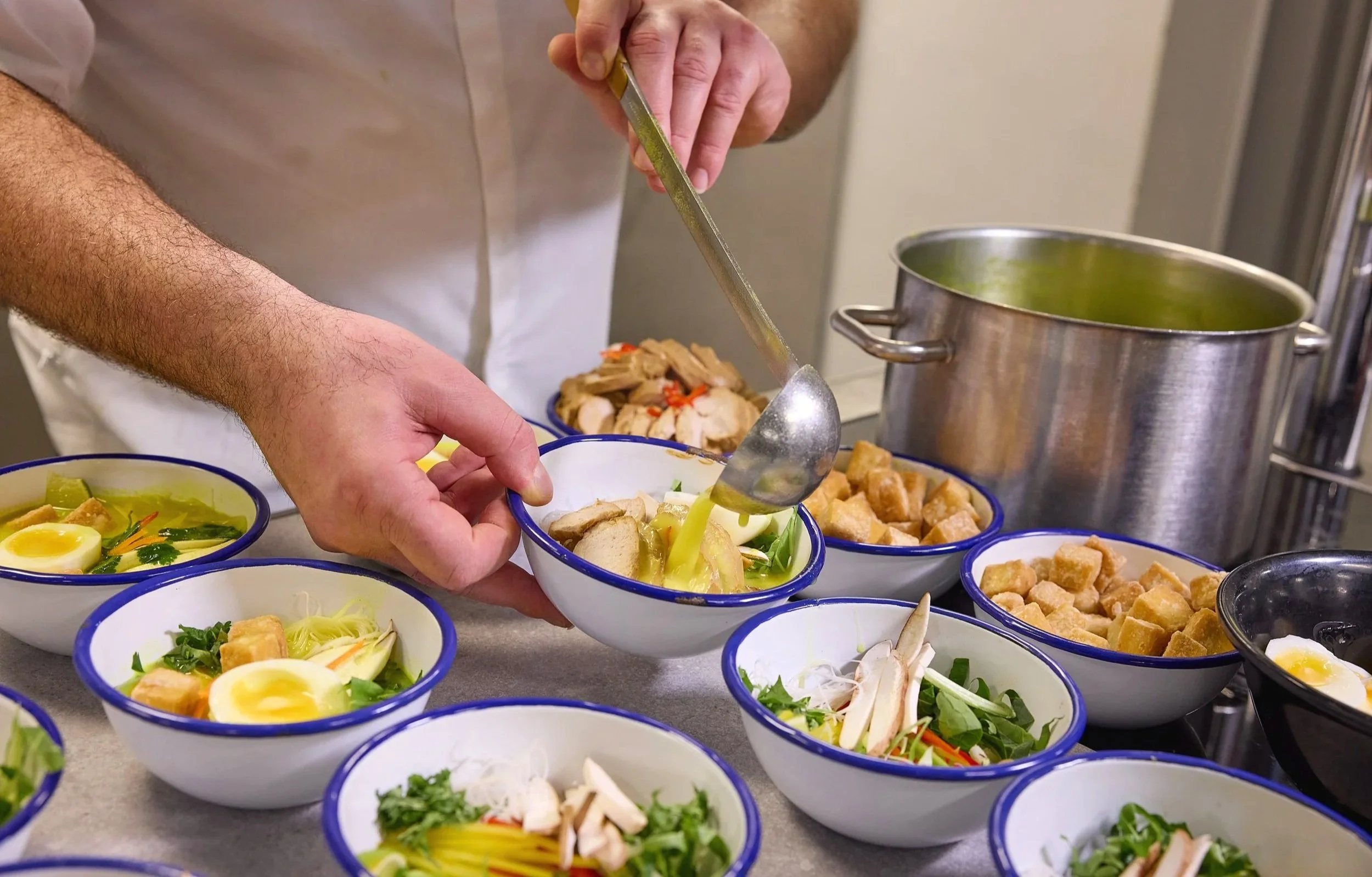A person serving food from a bowl into smaller bowls that contain salad ingredients such as lettuce, boiled eggs, and vegetables, with a pot of green soup nearby.