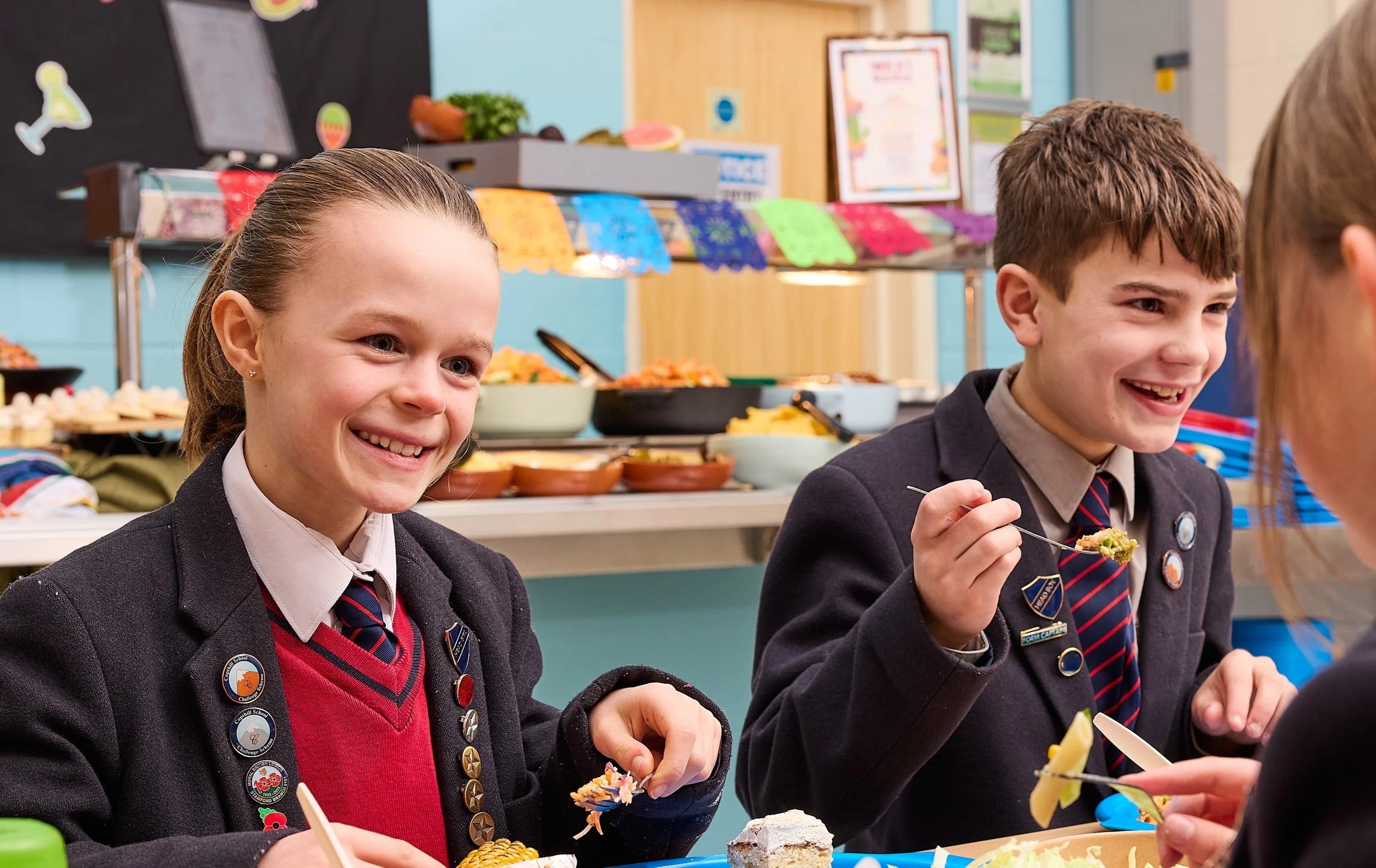 Two students in school uniforms eating desserts at a school event with colorful decorations and food in the background.