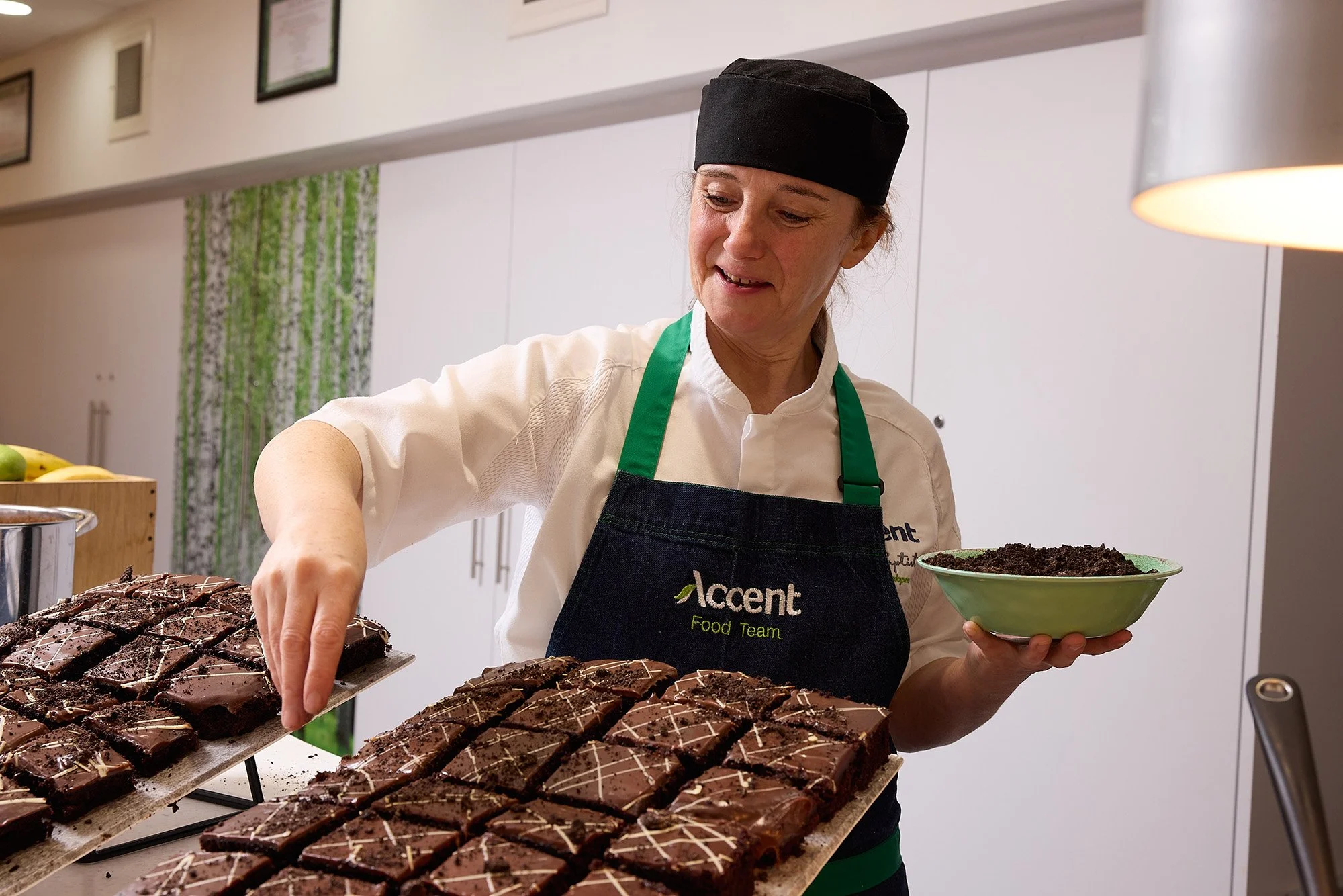 A woman wearing a black hat and apron, standing in a kitchen, is serving slices of chocolate cake from a tray onto a green plate.