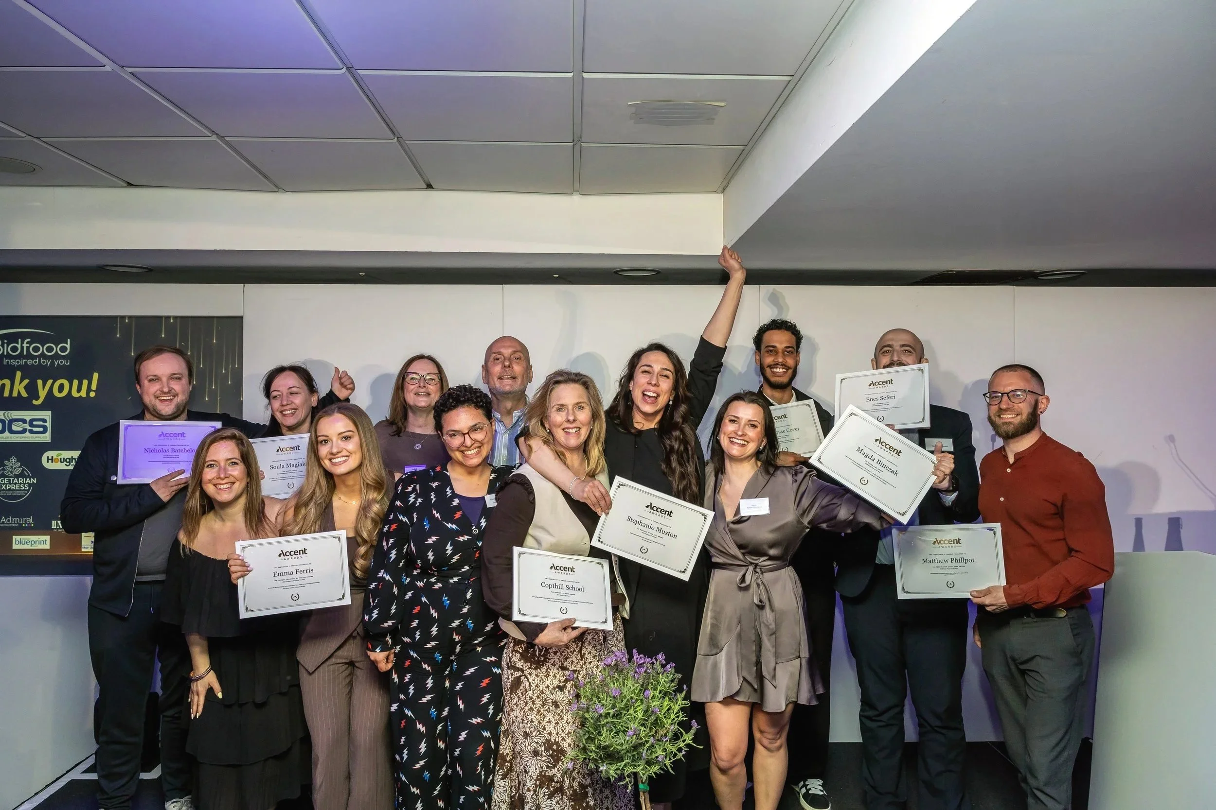 Group of smiling people holding certificates at an awards ceremony, standing in front of a dark backdrop with sponsor logos.