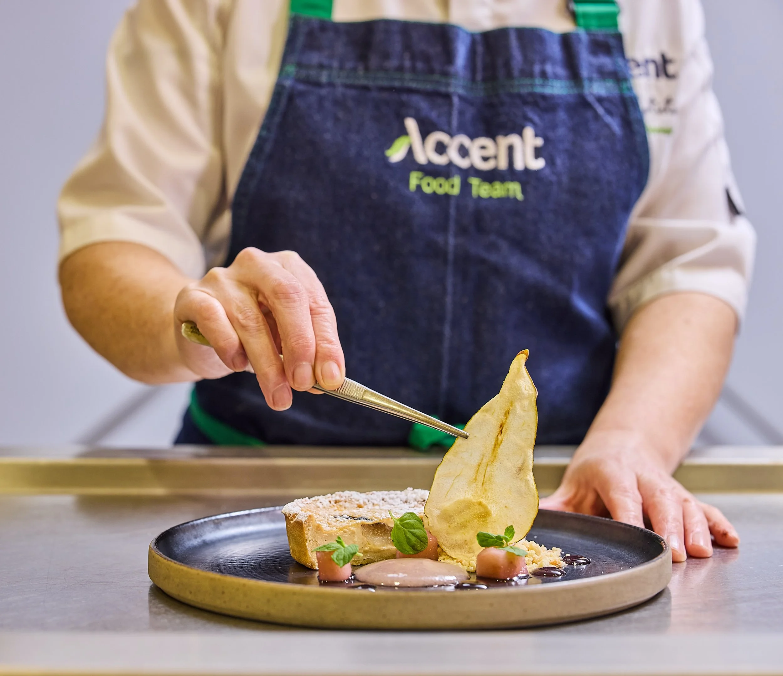 A chef in a green apron and denim bib apron with the words 'Accent Food Team' embroidered on it is garnishing a savory dish with potato chips and greens on a black plate.