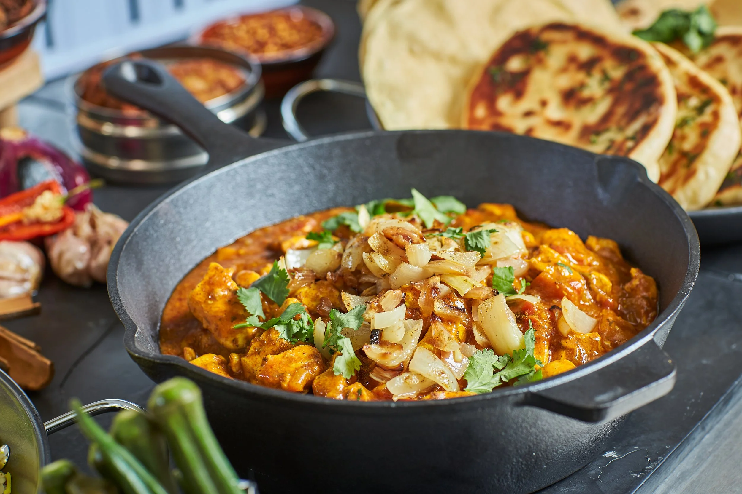 Close-up of a cast iron skillet with cooked chicken in a red sauce, topped with chopped onions and cilantro, on a black surface with Indian naan bread and small bowls of spices in the background.