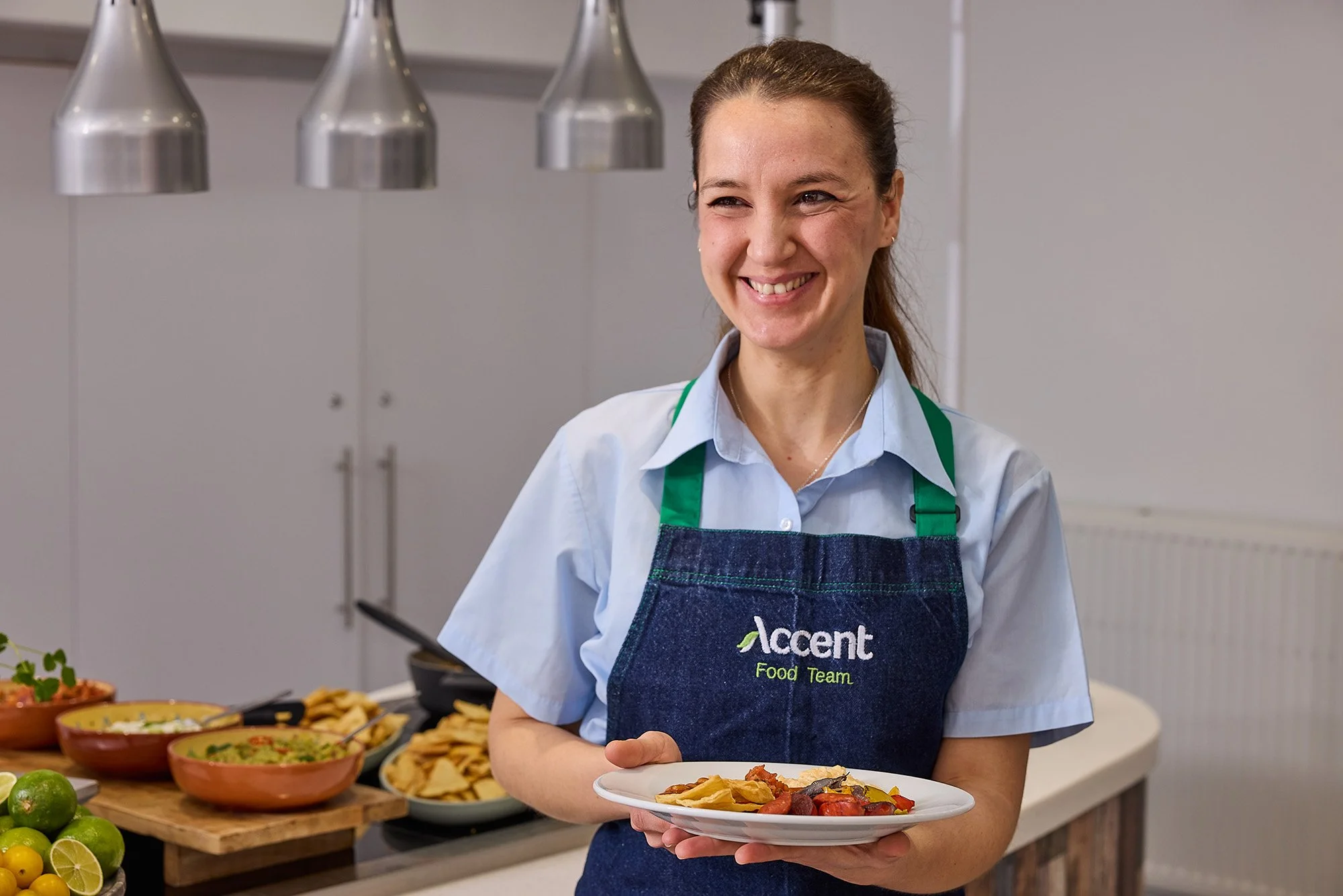 A smiling woman wearing an apron with 'Accenture Food Team' written on it, holding a plate of food, standing in front of a table with bowls of various dishes, in a kitchen or cafeteria setting.