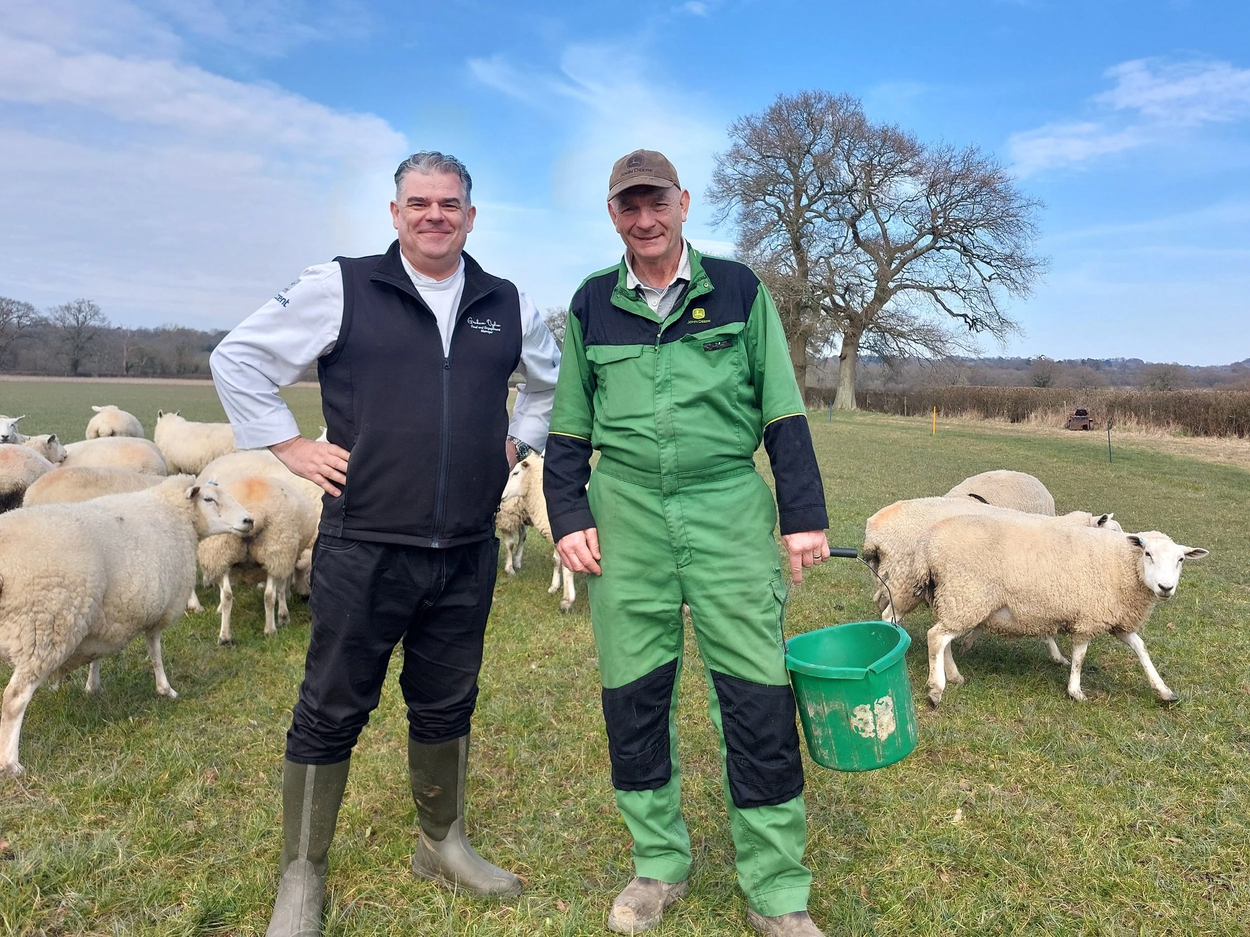 Two men stand outdoors on a farm with sheep, trees, and a blue sky in the background. One man has gray hair and is wearing a black vest over a white shirt, while the other is wearing a green and black jacket and a brown cap. They are smiling at the camera.