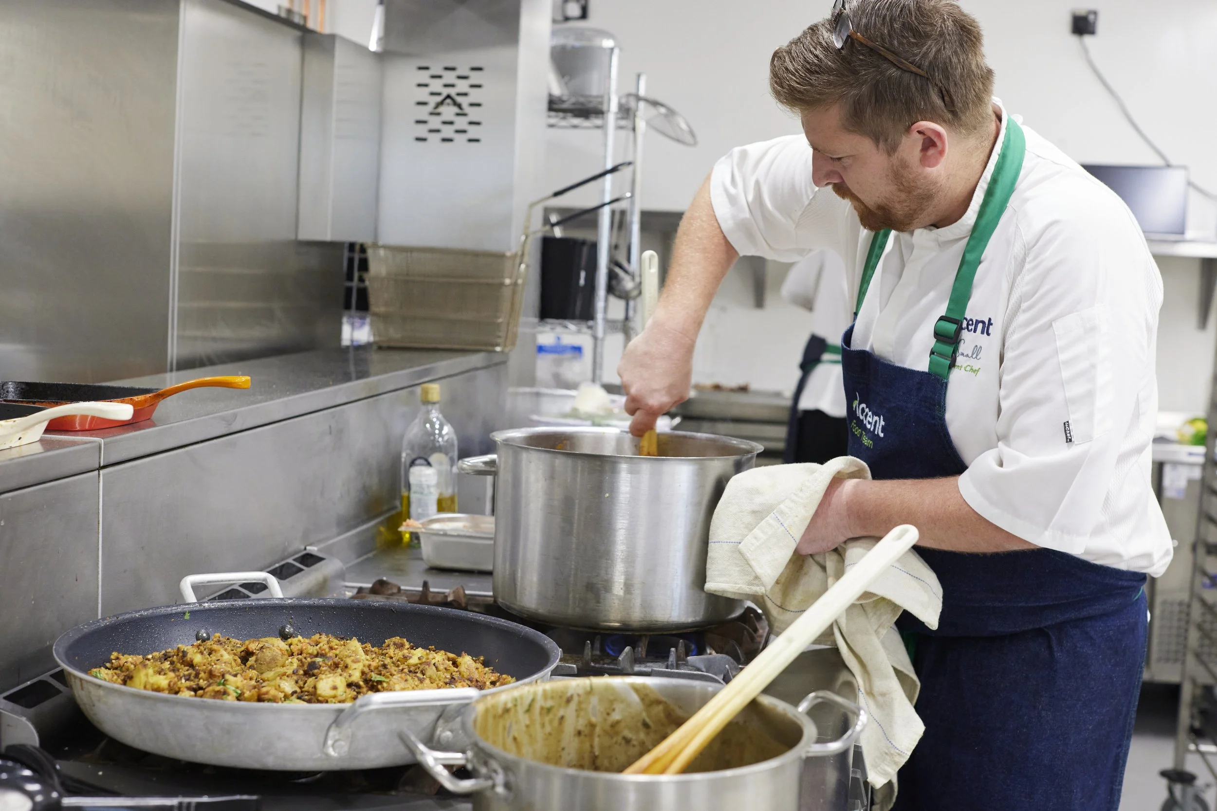 A chef cooking in a commercial kitchen, stirring a large pot with a beige towel, with various cooked dishes and cooking utensils around him.