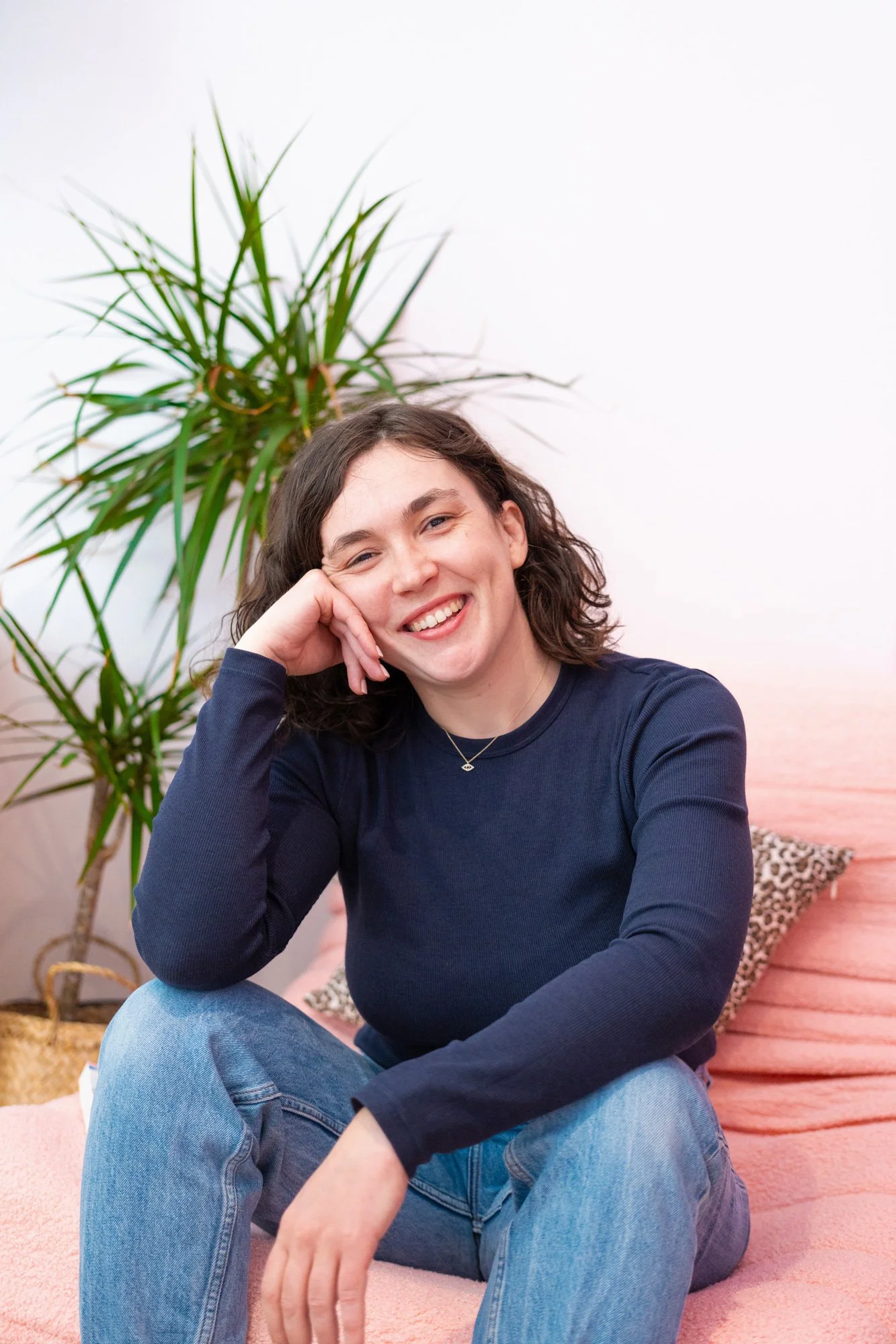 Young woman with dark wavy hair smiling while sitting on a pink carpet, resting her head on her hand, with a large green potted plant and a pink backdrop in the background.