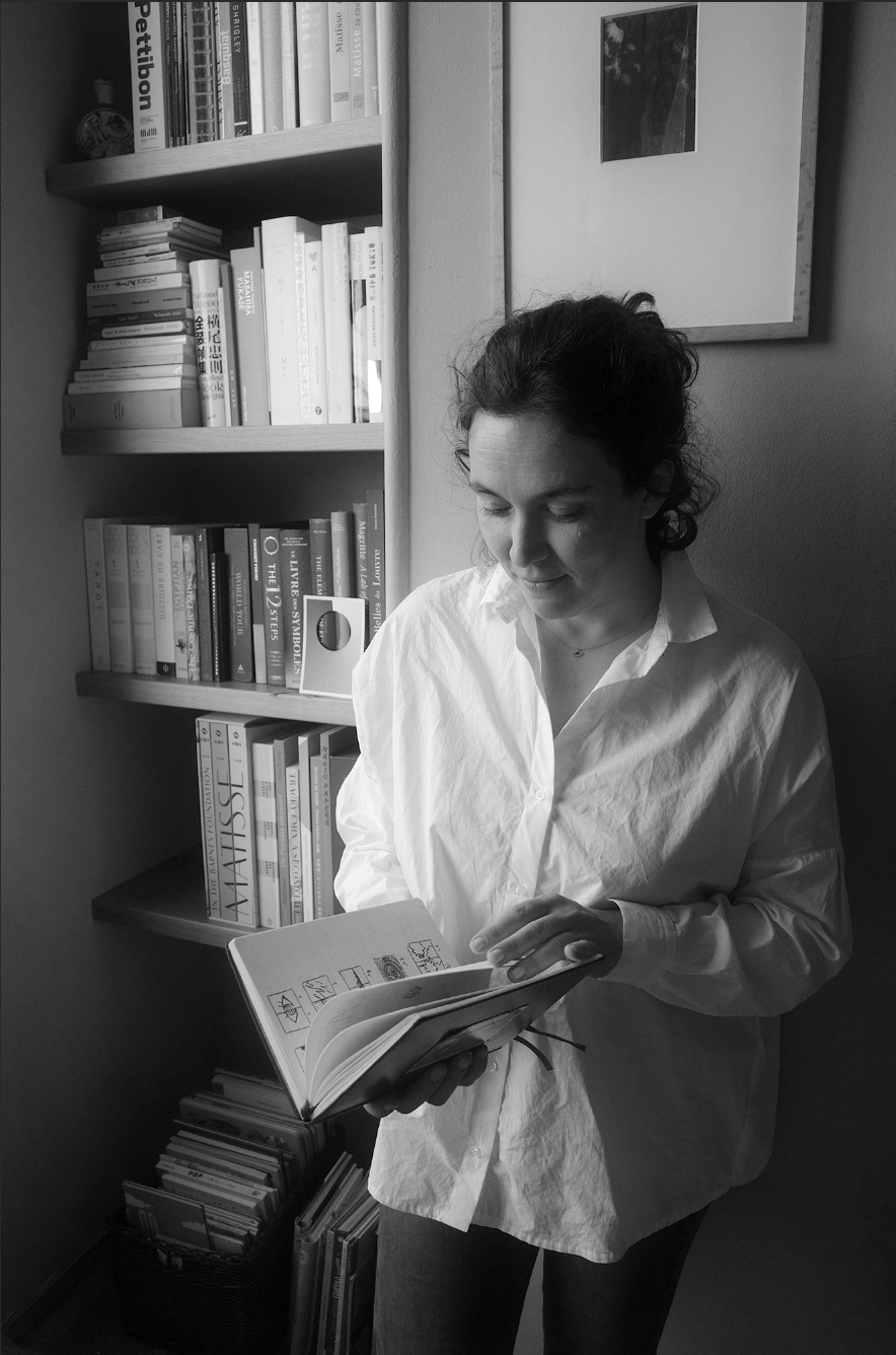 A woman with dark curly hair wearing a white button-up shirt, standing beside a bookshelf, reading an open book with illustrations, in a room with a framed picture on the wall.