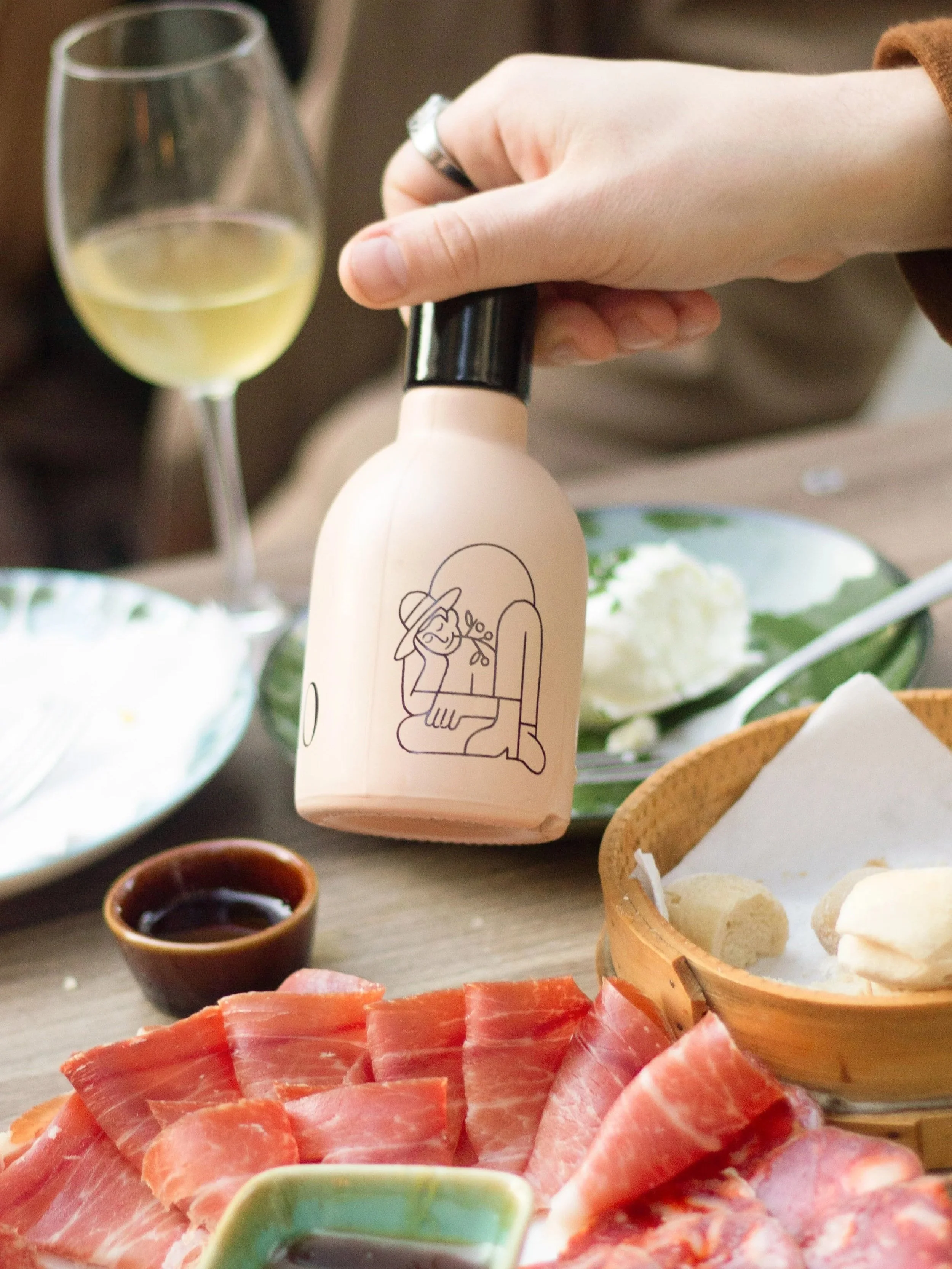 Pessoa segurando uma garrafinha de leite ou algum produto semelhante, com uma ilustração de uma mulher de chapéu na embalagem. Ao fundo, há uma taça de vinho branco e uma mesa com várias comidas, incluindo presunto, queijo, molho e pão.
