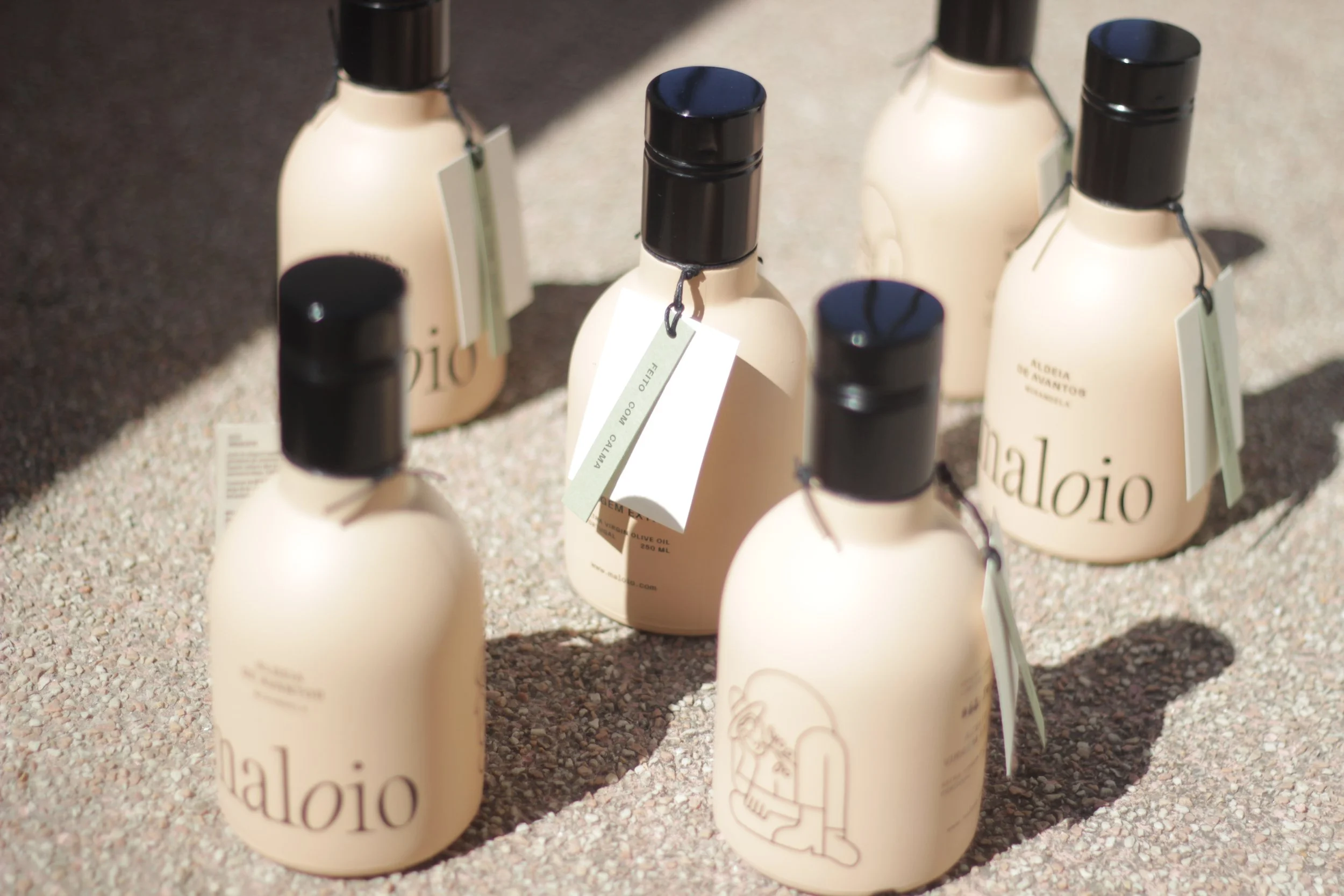 Garrafas de azeite de oliva com rótulos brancos e tampas pretas, colocadas sobre uma superfície de areia ou cascalho, com etiquetas penduradas.