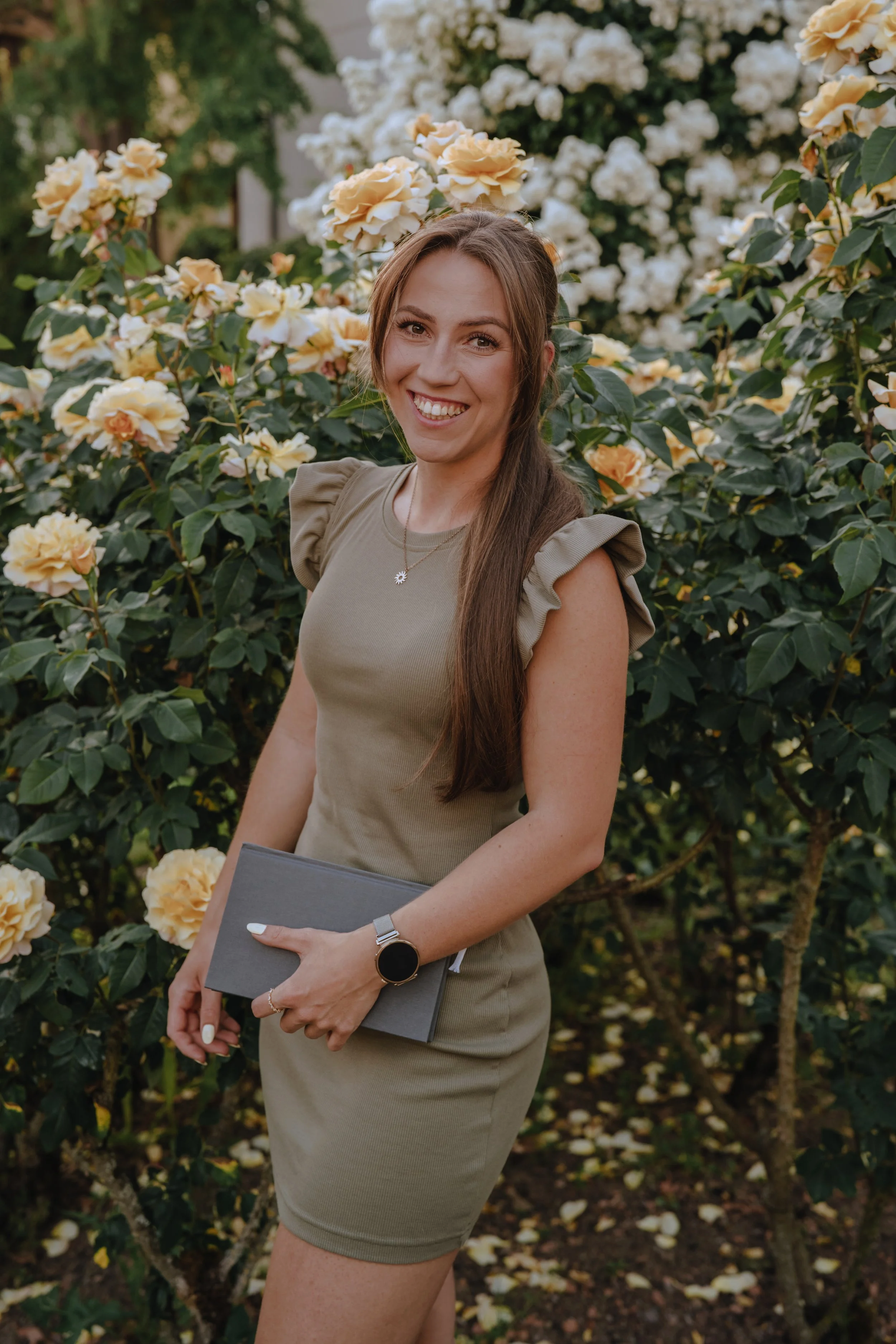 Eine lachende Frau steht vor einem Busch voller gelber und weißen Rosen, hält ein Buch oder einen Ordner in den Händen und trägt eine beige Bluse mit Raffung an den Schultern.