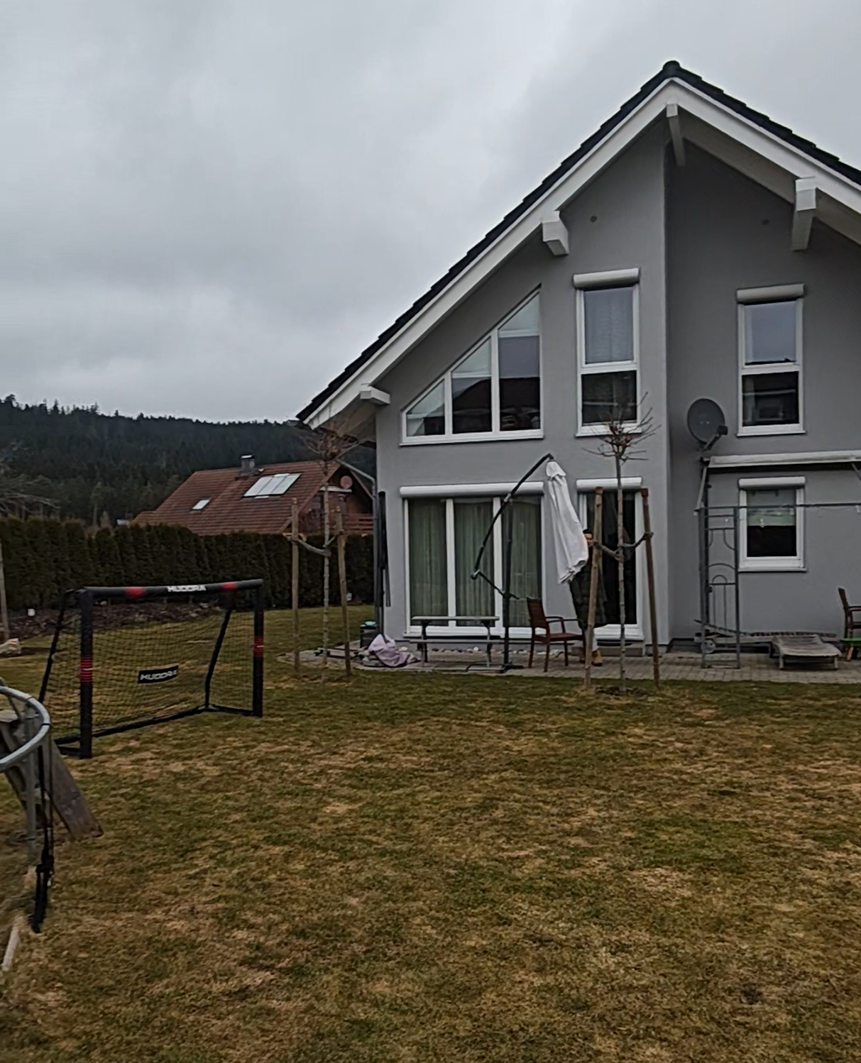 Ein zweistöckiges Haus mit grauer Fassade und großen Fenstern, im Garten befinden sich Spielsachen, ein Telt, Stühle, ein Sonnenschirm und Kinderfahrrad, im Hintergrund Bäume und andere Häuser bei bewölktem Himmel.